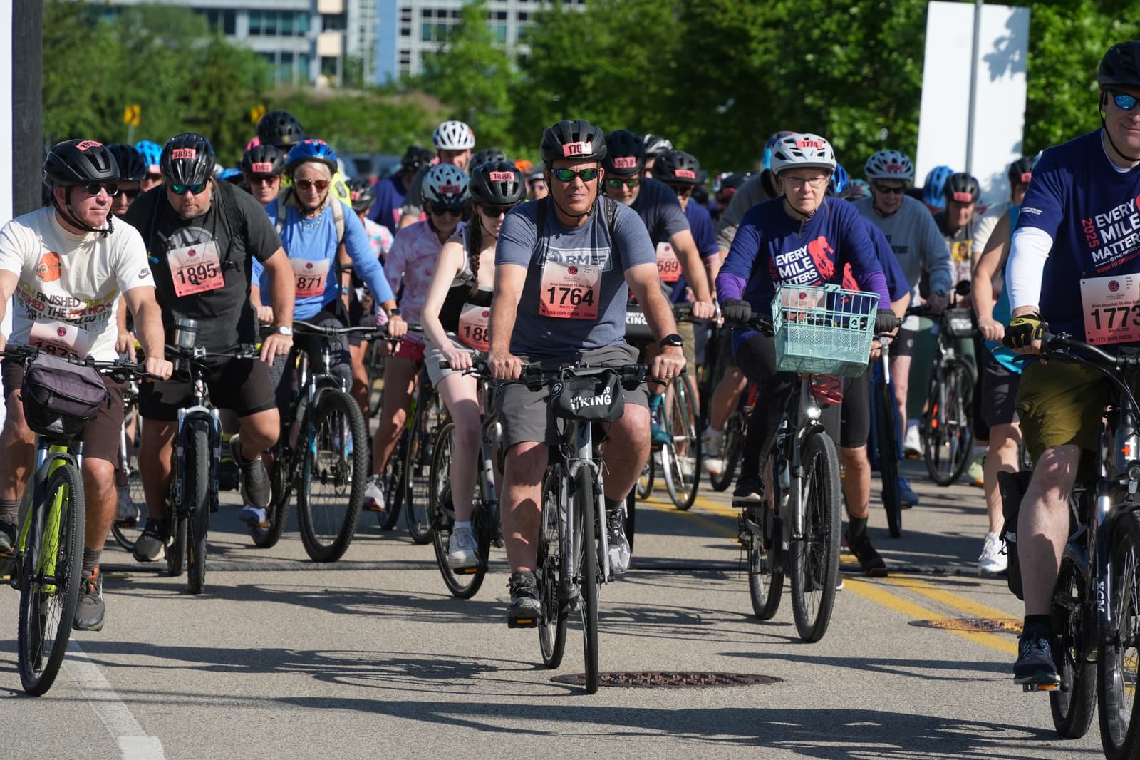 A group of cyclists wearing helmets and race numbers ride together on a sunny day, participating in an organized cycling event on a paved road. Trees and buildings are visible in the background.
