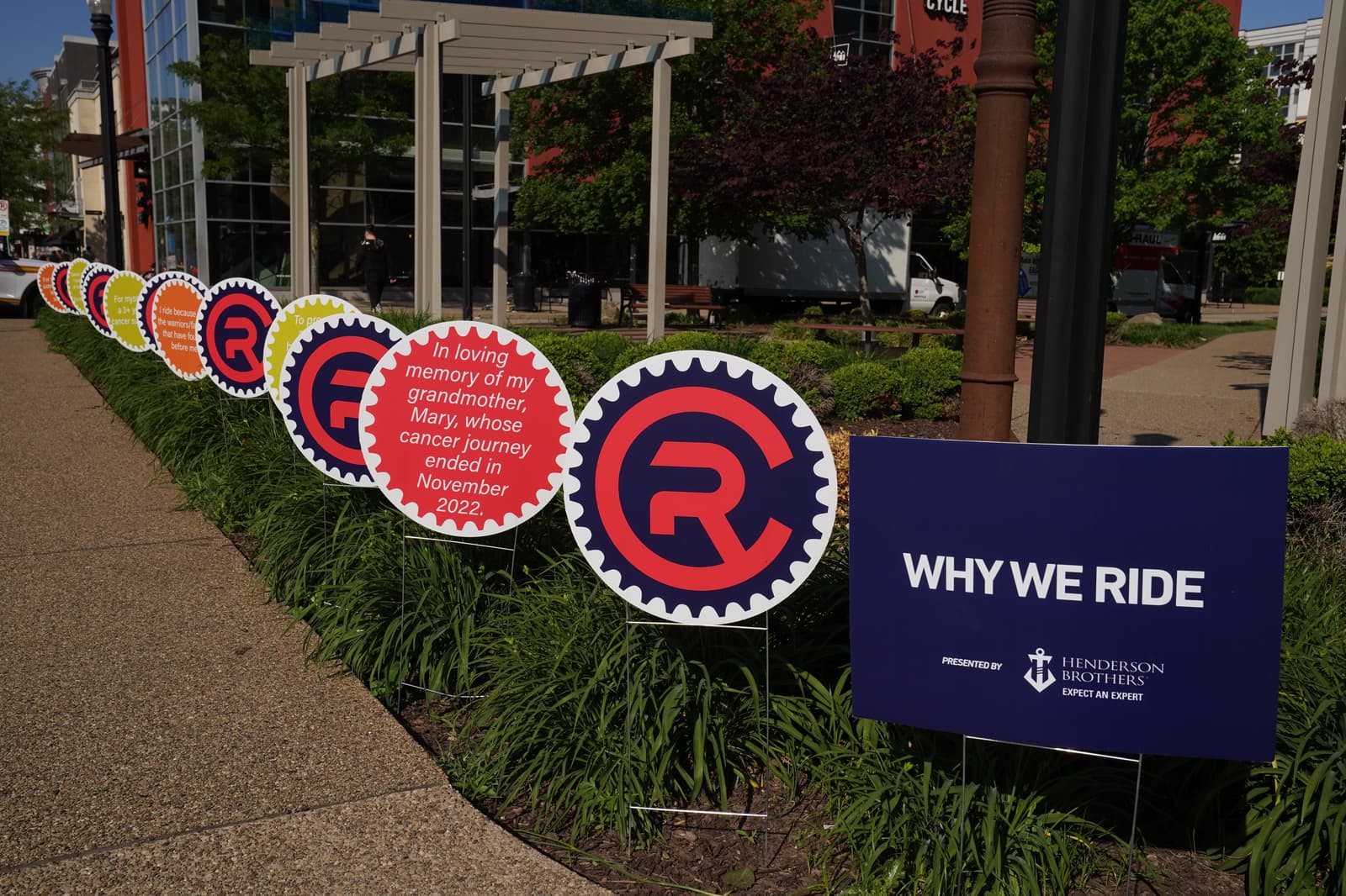 Colorful circular signs line a garden bed, one reading "In loving memory of my grandmother, Mary, whose cancer journey ended in November 2022." A blue sign says "WHY WE RIDE" near a building and shaded sidewalk.