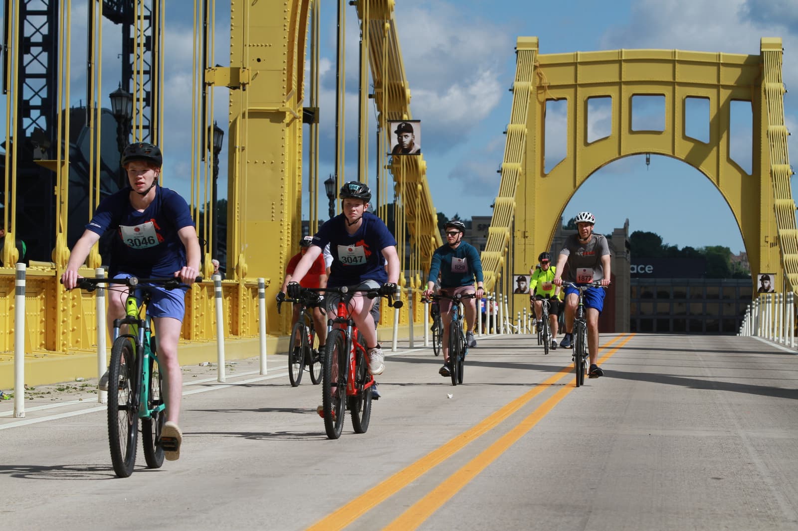 A group of cyclists wearing helmets and race bibs ride across a bright yellow bridge on a sunny day, with blue skies and scattered clouds in the background.