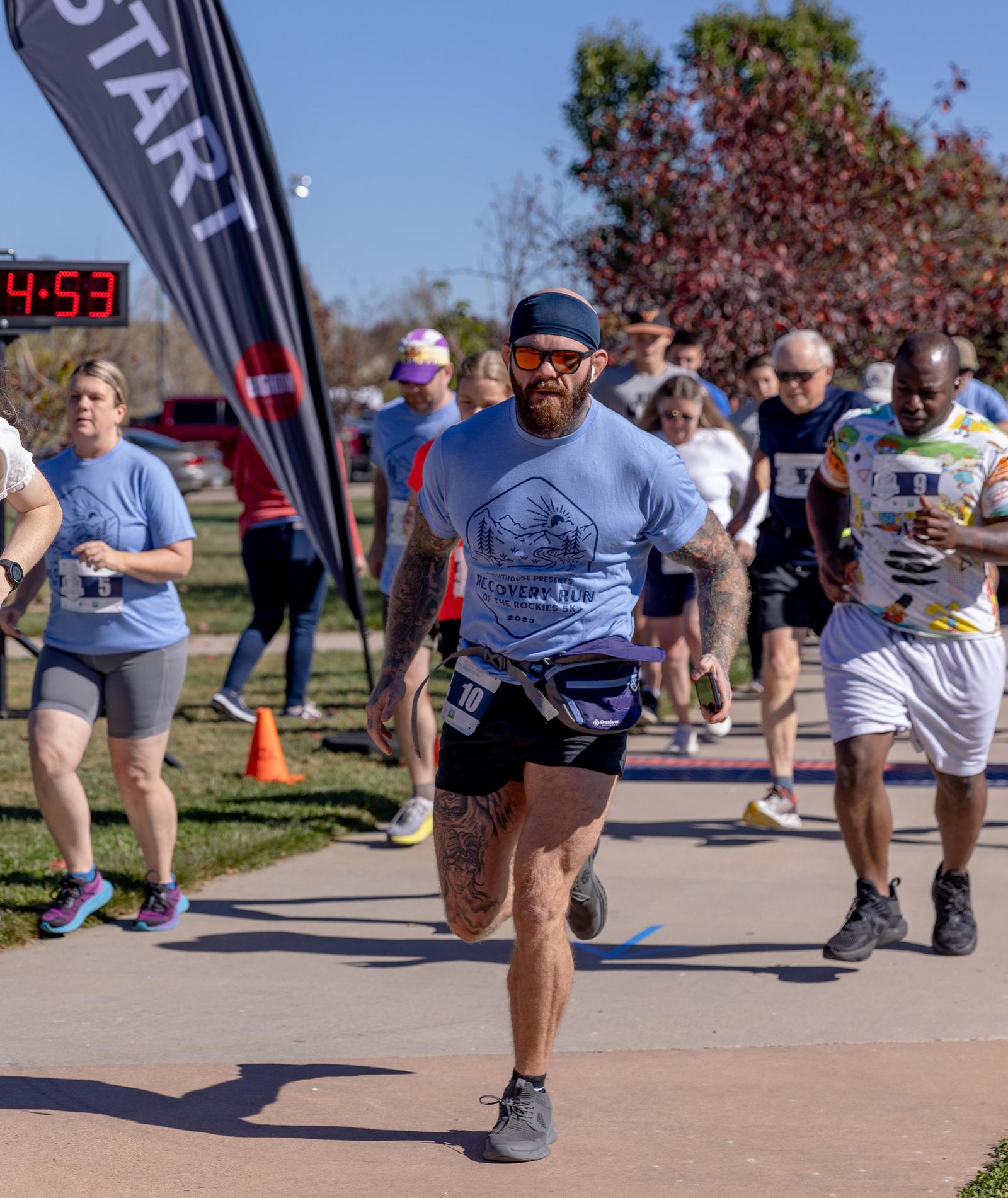 A group of runners, including a bearded man with tattoos and sunglasses in front, cross the starting line of a race on a sunny day. A digital clock and a "START" flag are visible in the background.