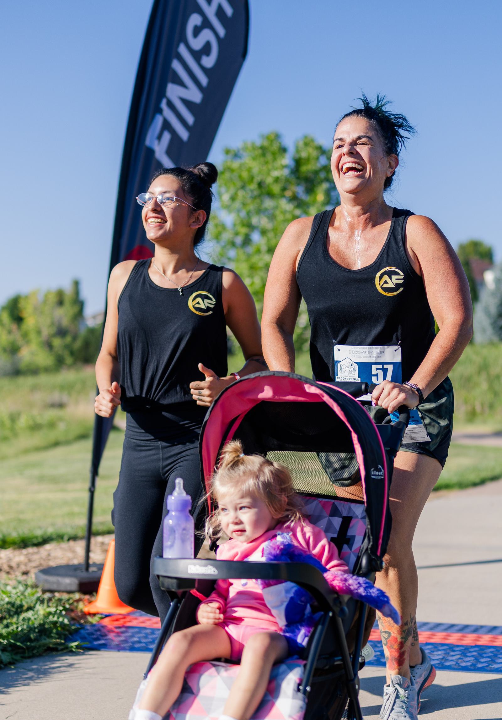 Two women wearing matching black athletic tops run and smile near a "Finish" banner while pushing a young girl in a stroller on a sunny outdoor path. The girl looks ahead, holding a bottle.
