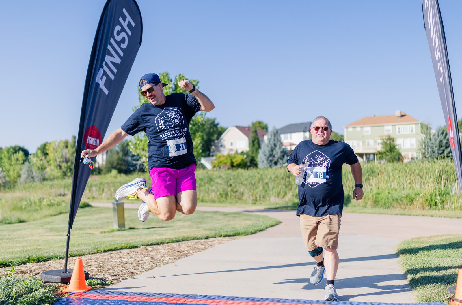 Two men with race bibs, one jumping joyfully and the other smiling, cross a finish line outdoors on a sunny day. They are wearing matching shirts and running gear, with greenery and houses in the background.