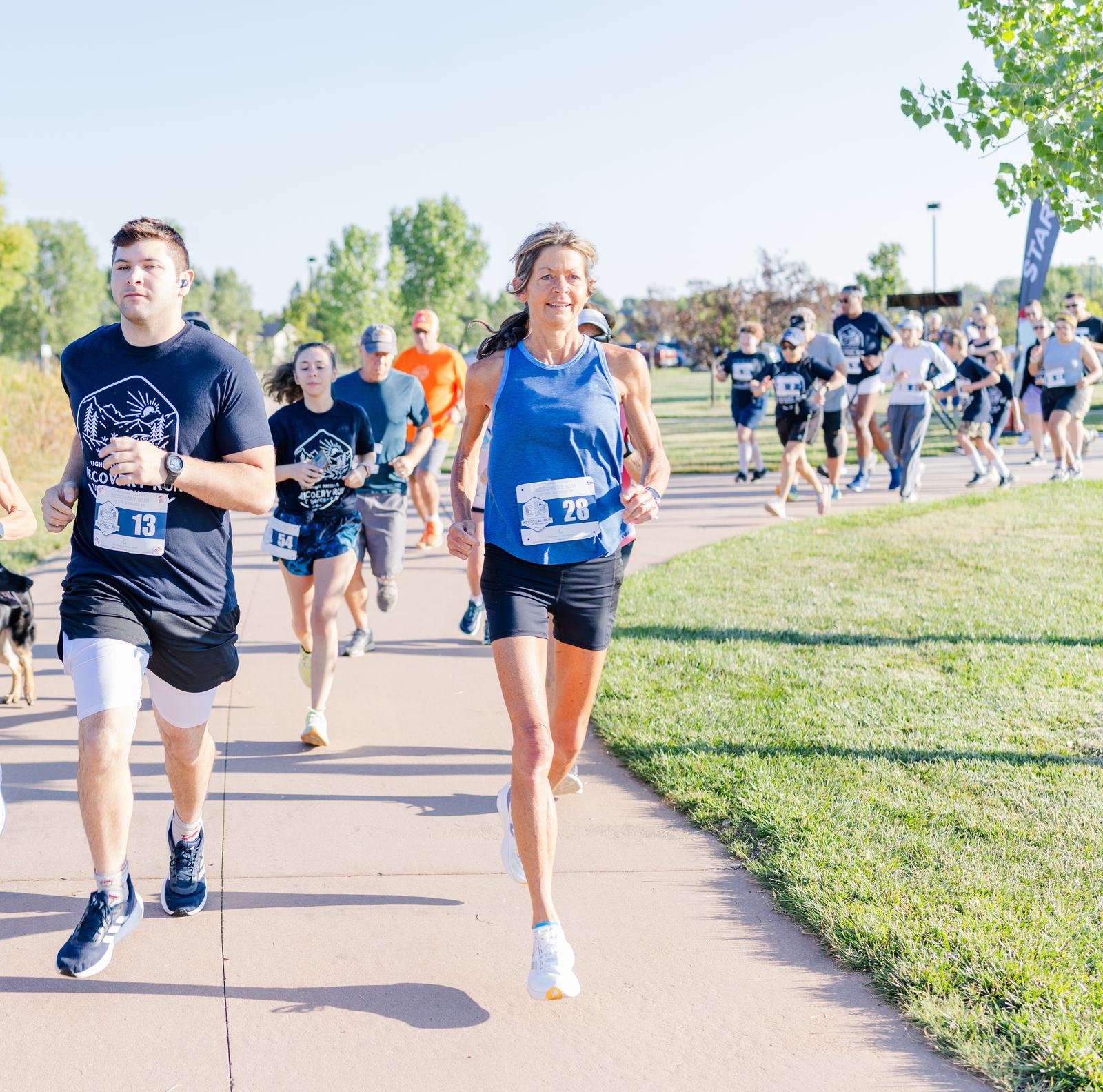 A group of runners, each wearing race bibs, jog along a paved park path surrounded by greenery on a sunny day. The participants look focused and determined.
