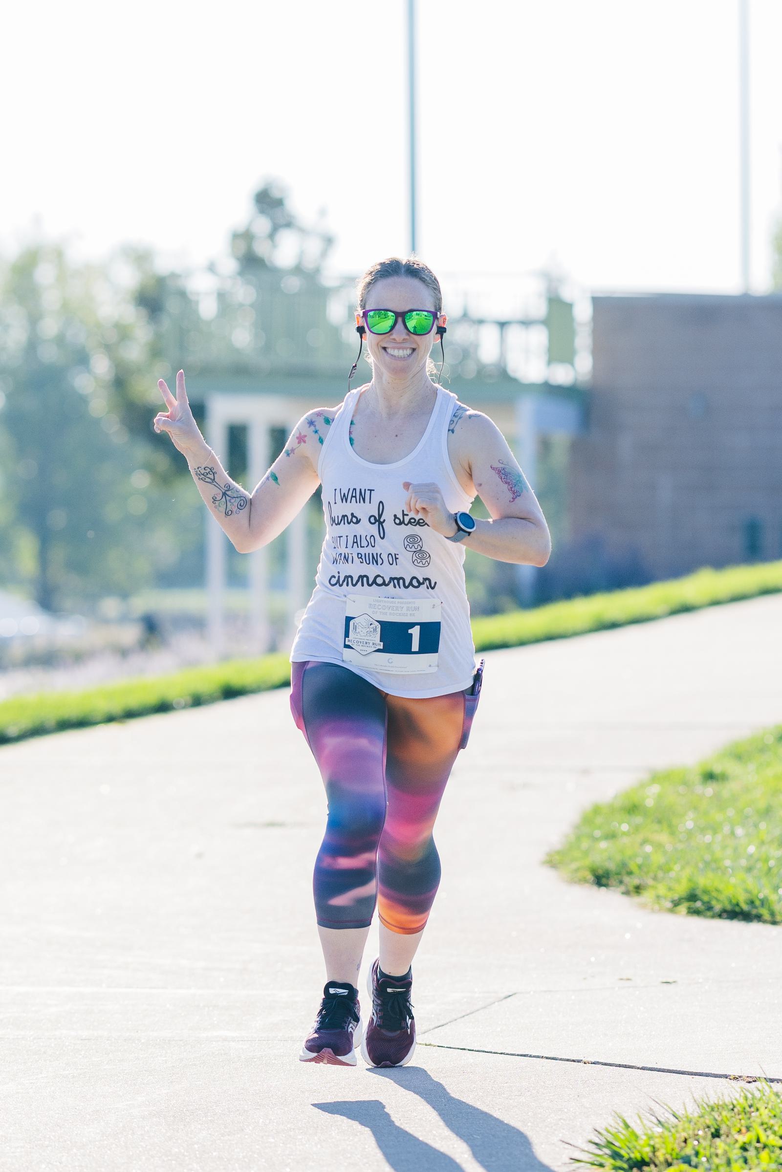 A woman wearing sunglasses, colorful leggings, and a tank top making a peace sign while jogging on a sunny outdoor path. She has a race bib with the number 1 and is smiling enthusiastically.