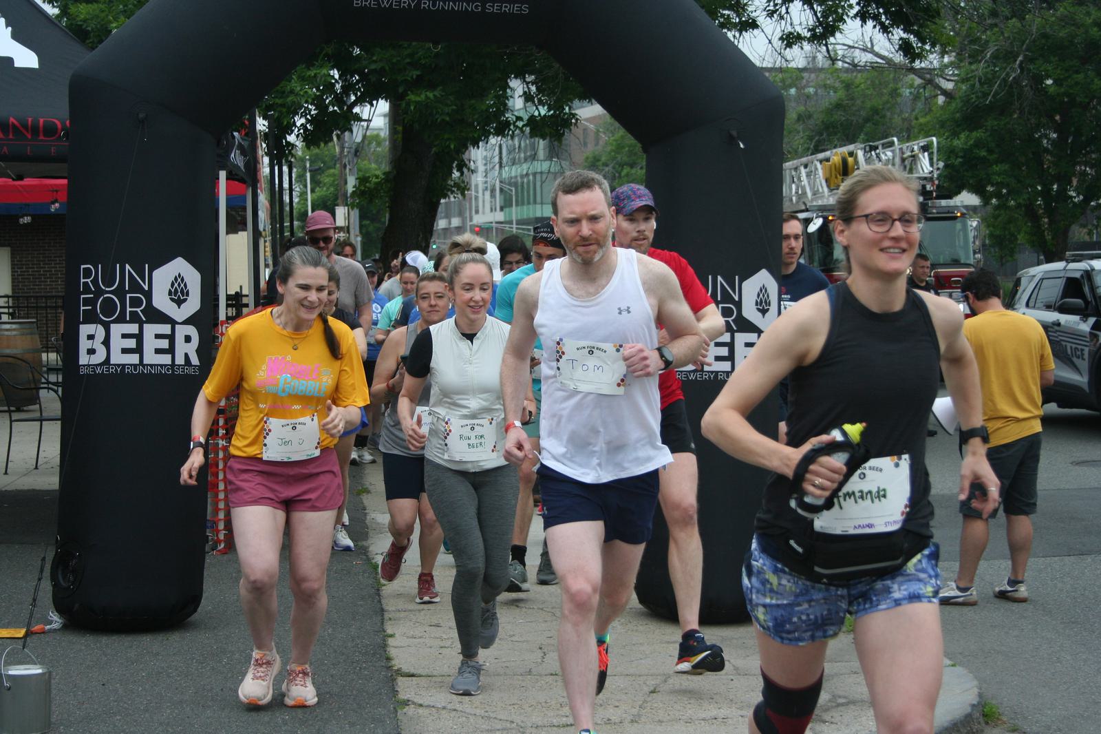 A group of runners, wearing race bibs, starts a run beneath an inflatable arch that reads "RUN FOR BEER." Participants appear cheerful and energetic as they begin the race on a city street.