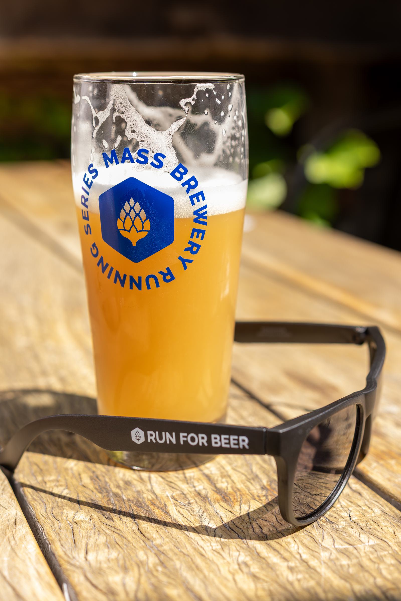A pint glass of hazy beer with "Mass Brewery Running Series" printed on it sits on a wooden table next to black sunglasses that read "Run For Beer." Sunlight creates bright highlights and shadows.