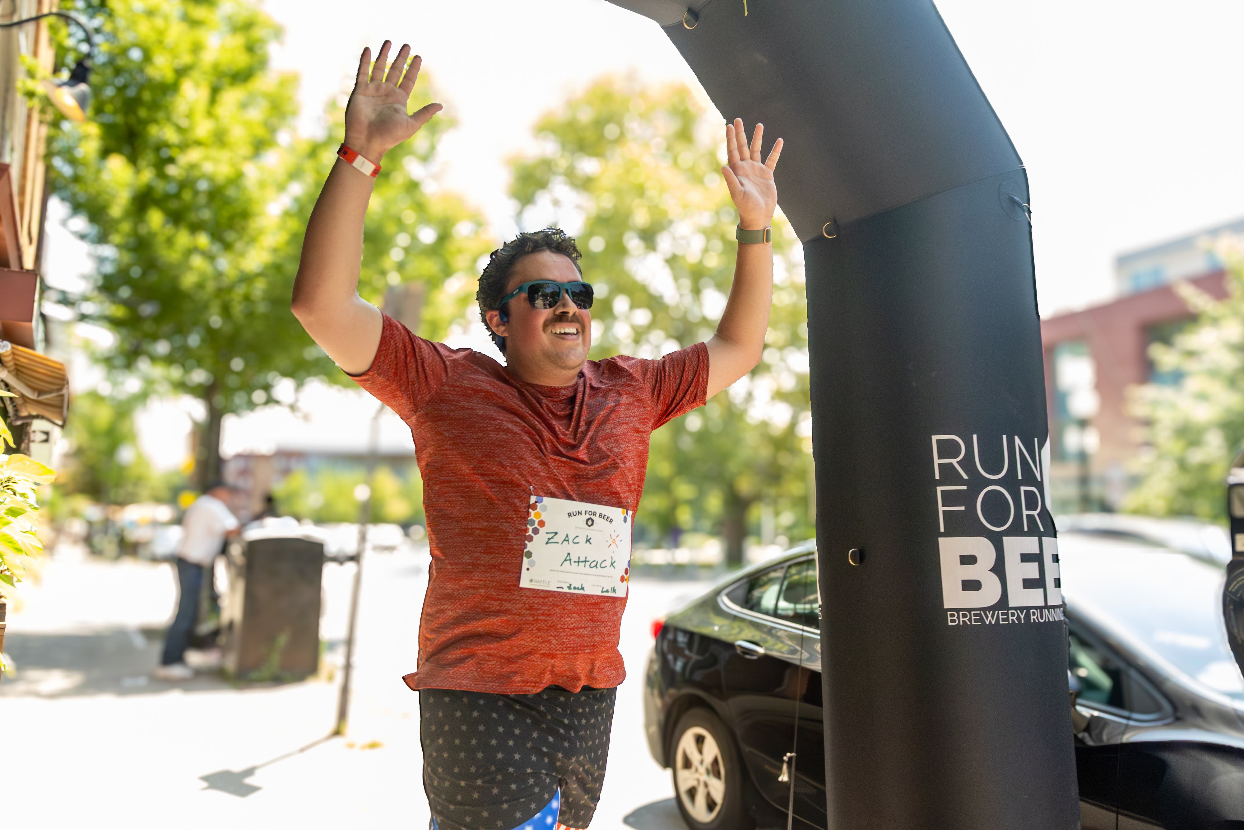 A smiling man in a red shirt and sunglasses raises his arms in celebration as he finishes a race. He wears a race bib and stands near a black archway labeled "RUN FOR BEER" on a sunny day.