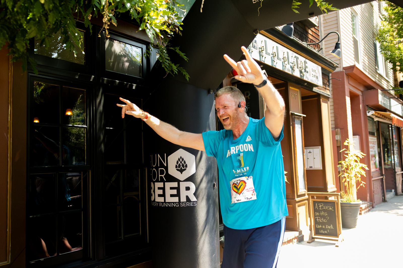 A man in a blue athletic shirt joyfully raises his arms and makes a hand gesture while running past a "Run for Beer" event sign on a sunny street lined with shops.