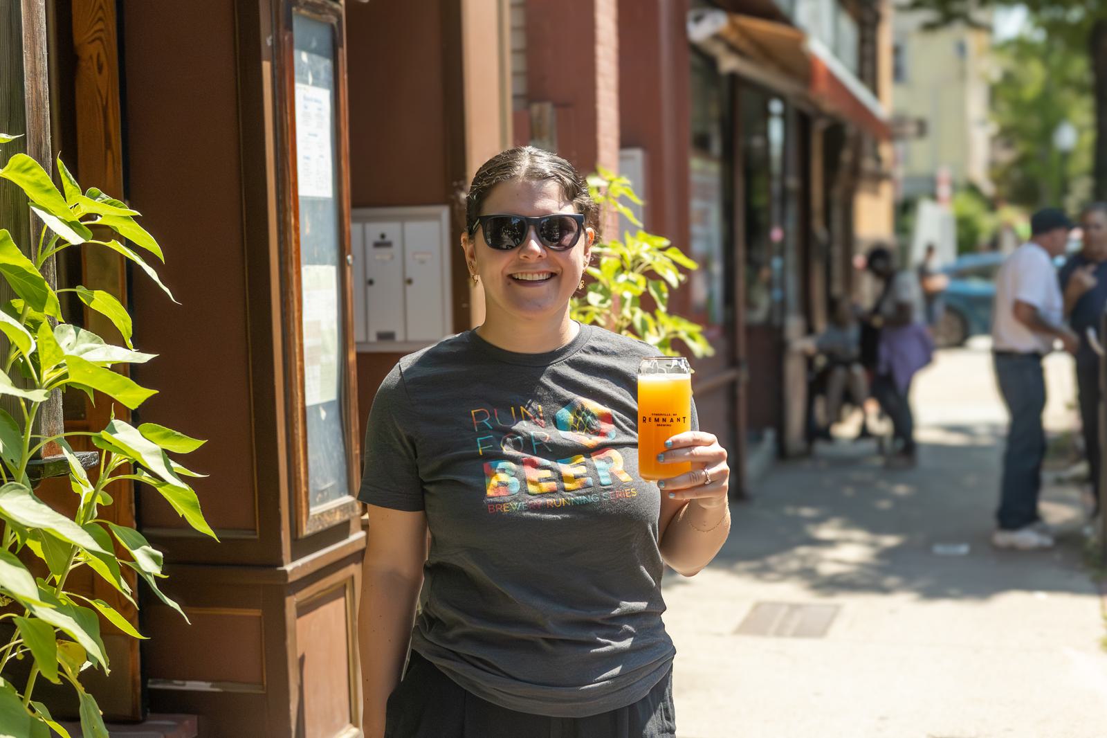 A smiling person wearing sunglasses and a “Run for Beer” shirt stands outside on a sunny day, holding a glass of beer. Green plants and storefronts line the sidewalk in the background.