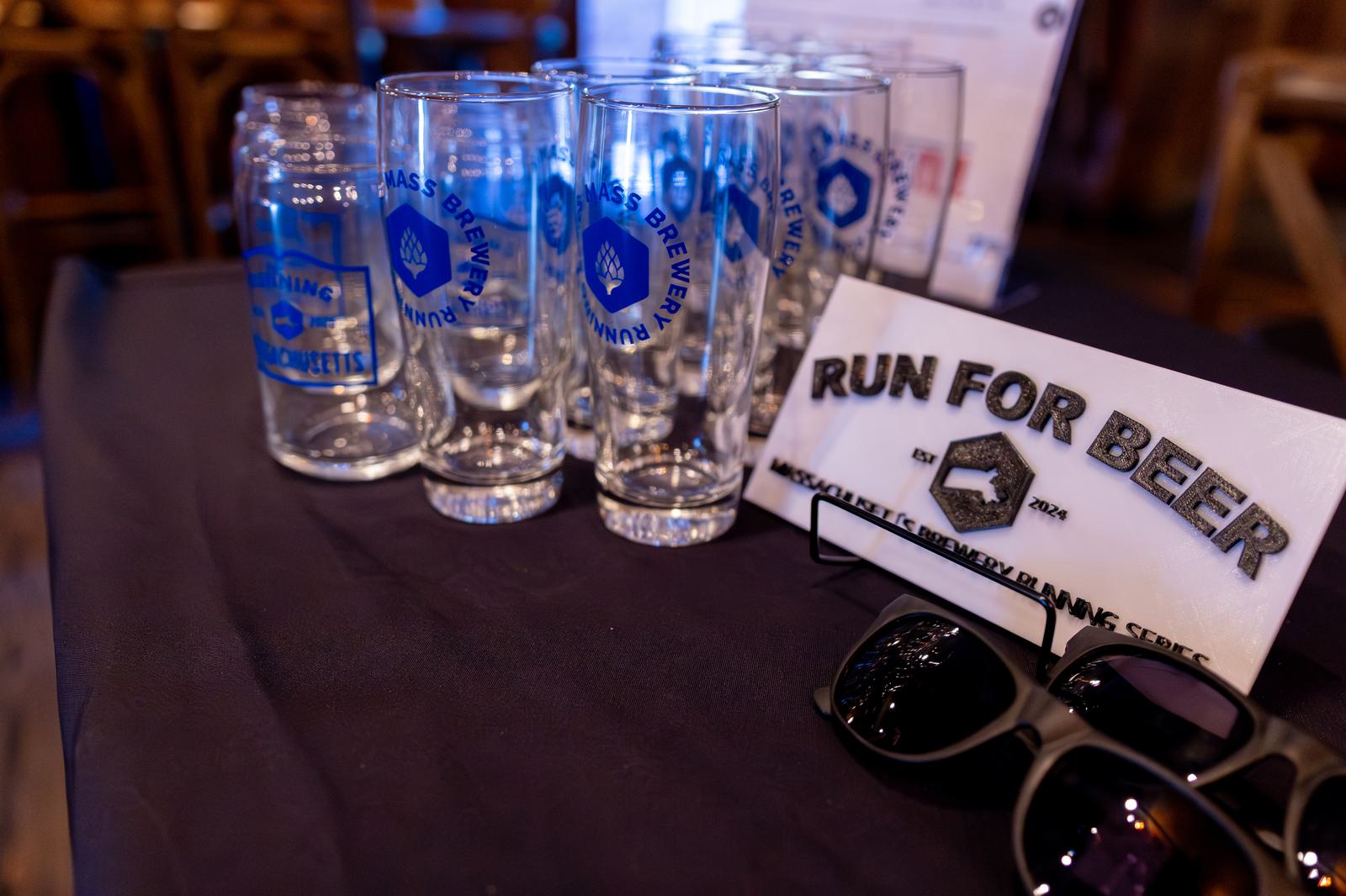 A group of clear beer glasses with blue logos is displayed next to a "RUN FOR BEER" sign and a pair of black sunglasses on a dark tablecloth.