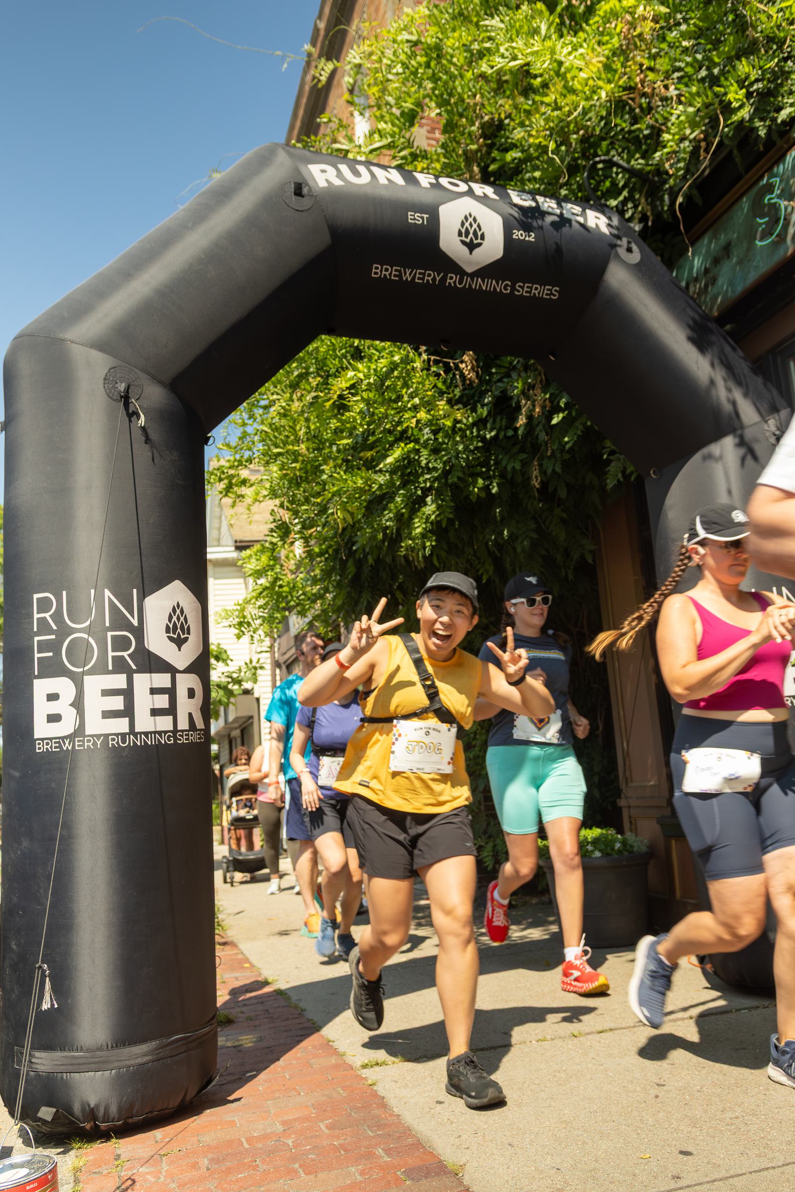 A group of runners smiles and waves as they pass under a black inflatable arch that reads "Run For Beer" during a sunny outdoor fun run event. The atmosphere is lively and energetic.