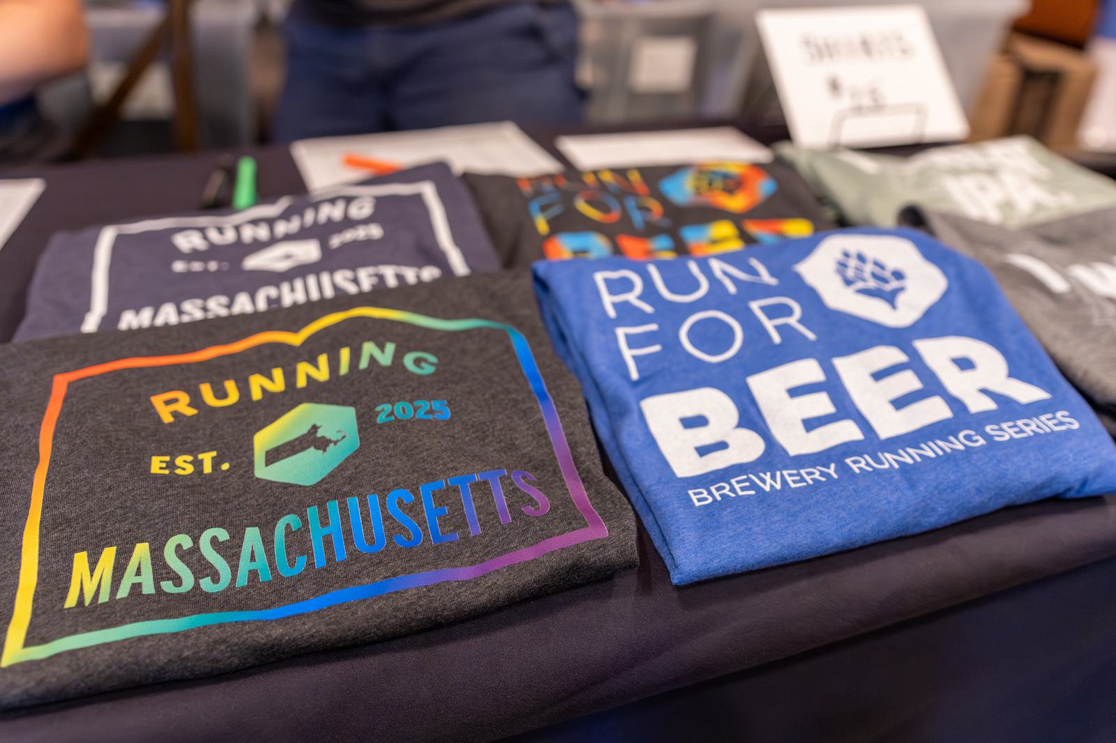 A table displays colorful running-themed T-shirts, including one reading "RUNNING MASSACHUSETTS Est. 2025" with a rainbow border, and another reading "RUN FOR BEER Brewery Running Series.