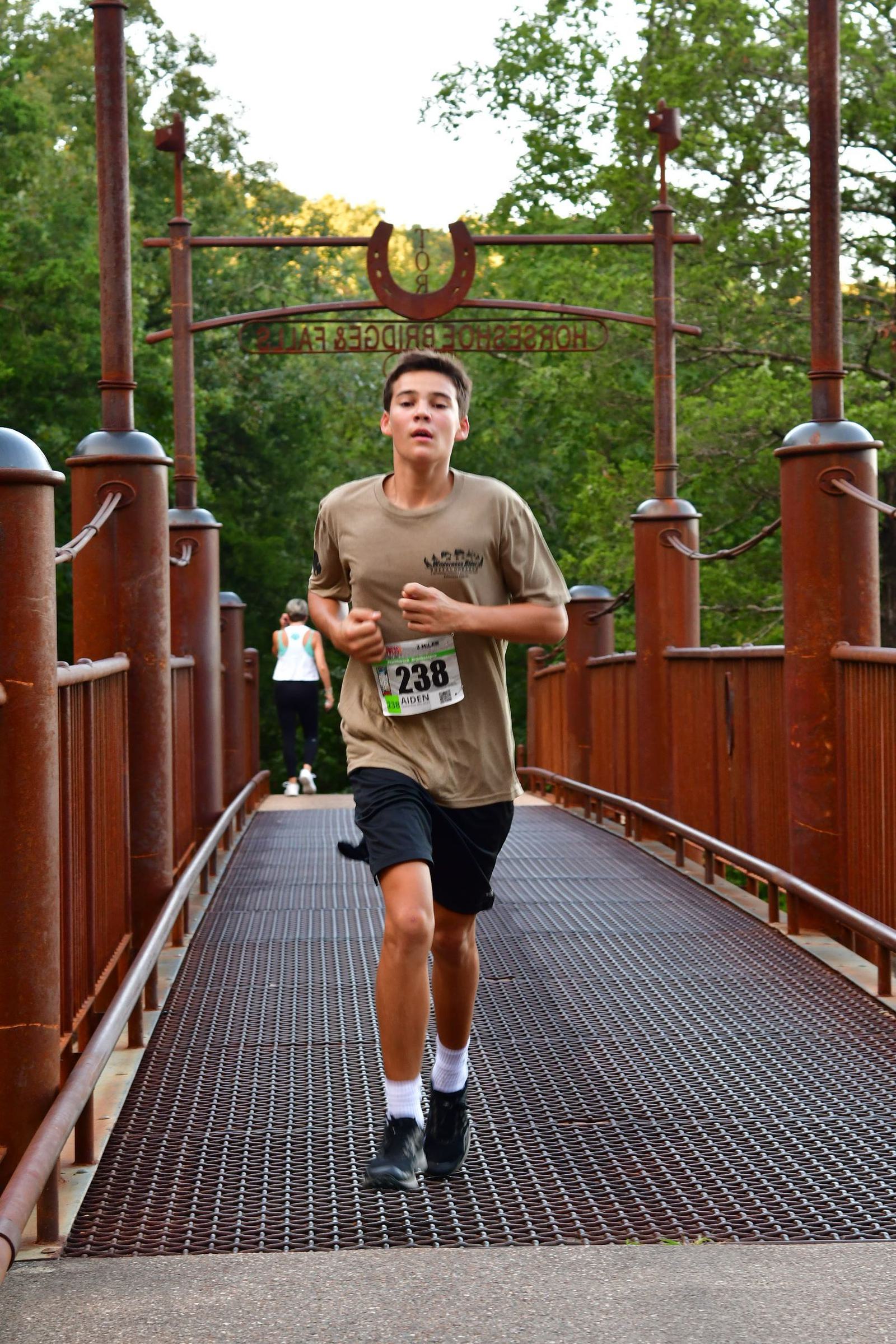 A young man wearing a brown T-shirt and black shorts runs across a brown metal bridge during a race. He has a race bib numbered 238. Green trees and a sign reading "Horsehoe Bridge" are visible in the background.