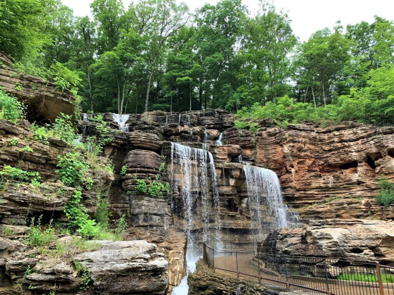 A multi-tiered waterfall cascades over layered rocky cliffs, surrounded by lush green trees. Metal railings and a walkway are visible at the base of the falls.