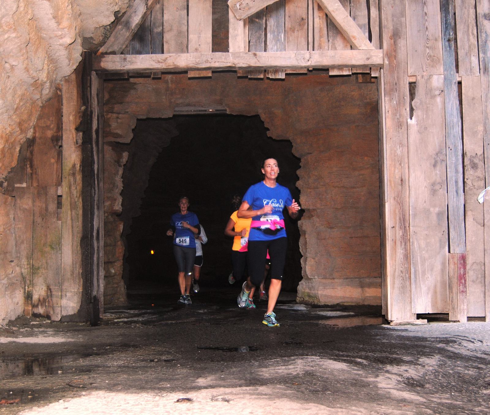 Runners emerge from a dark, rocky tunnel with a wooden entrance, participating in a race. The lead runner wears a blue shirt and black pants, followed by others in athletic gear.