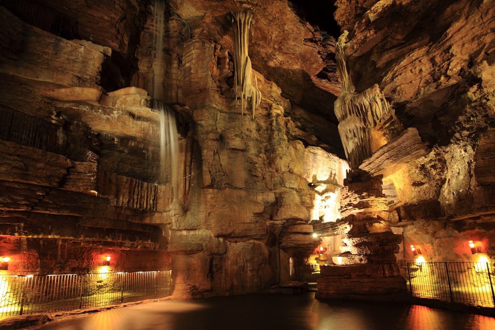 A large, illuminated underground cave with rugged rock walls, stalactites hanging from the ceiling, a small waterfall, and a pool of water, with metal railings and soft lighting along the paths.