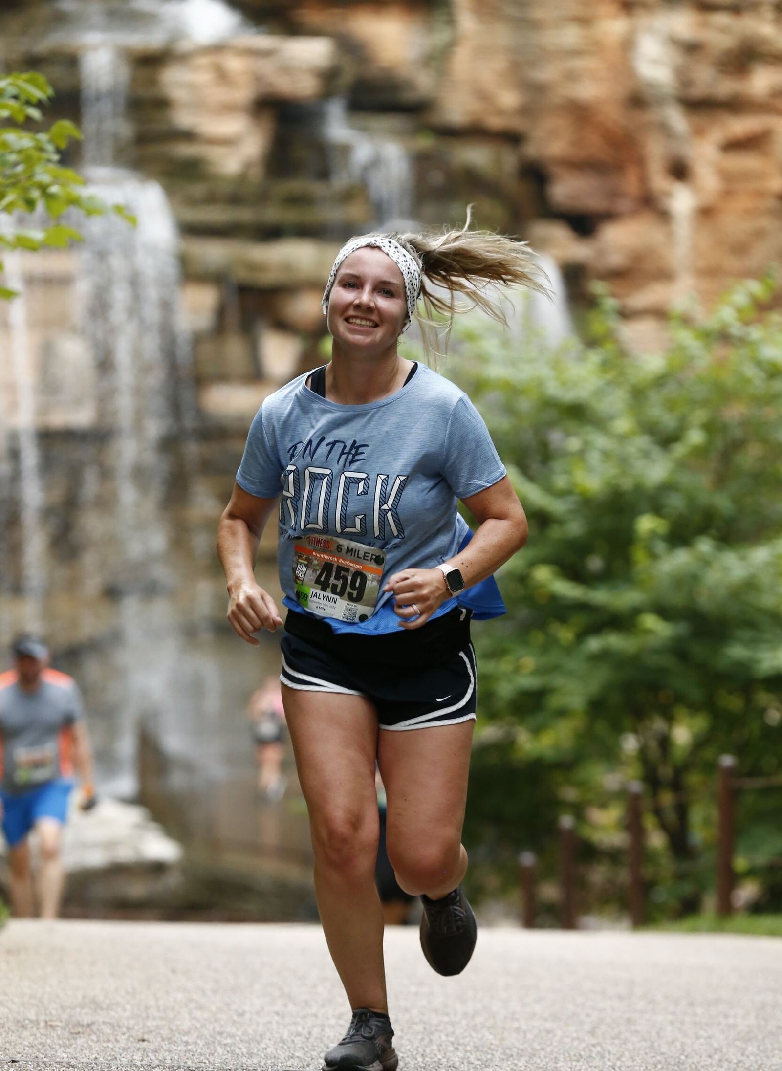 A smiling woman runs outdoors in a race, wearing a gray “On the Rock” shirt, black shorts, and a race bib. Waterfalls and greenery are visible in the background.