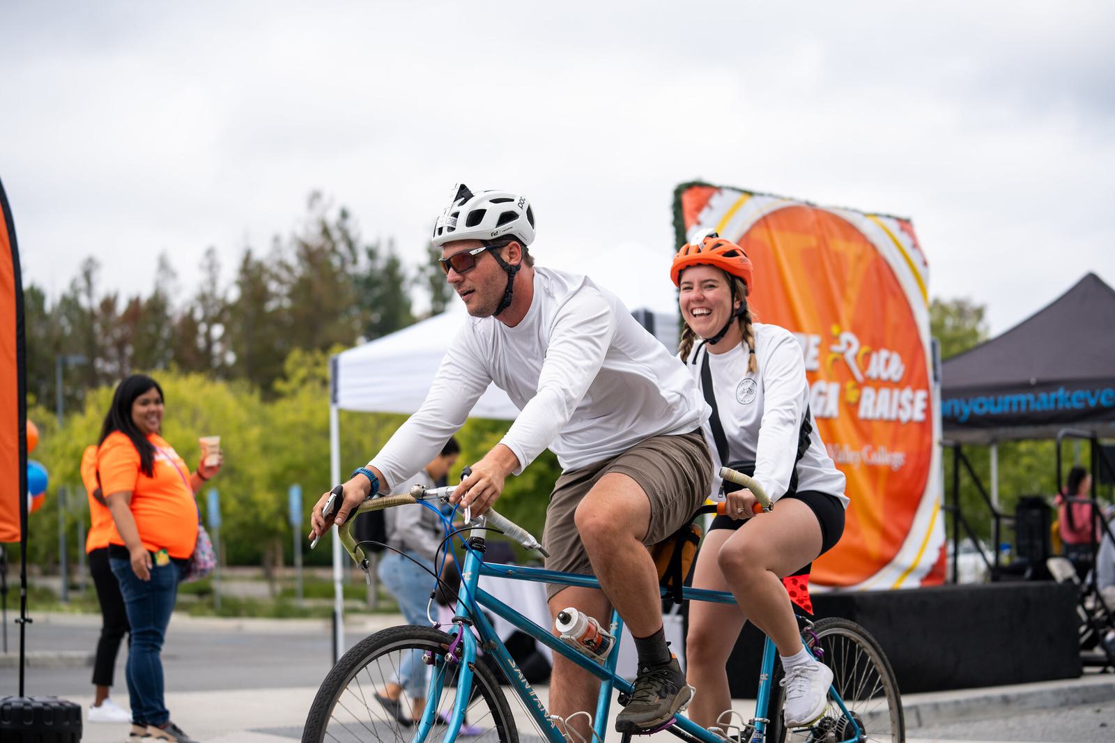 Two people wearing helmets and white shirts ride a tandem bicycle, smiling. An orange event banner, tents, and a few other people are visible in the background on a cloudy day.