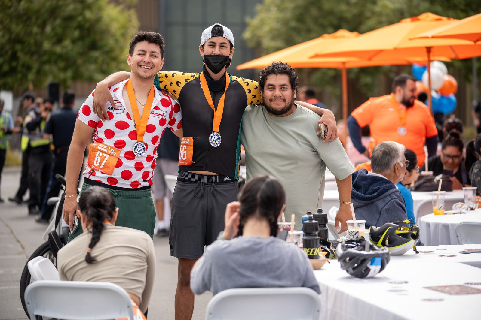 Three men pose and smile with their arms around each other at an outdoor event. Two wear medals and race bibs. People sit at tables with food and drinks. Orange umbrellas and balloons decorate the background.