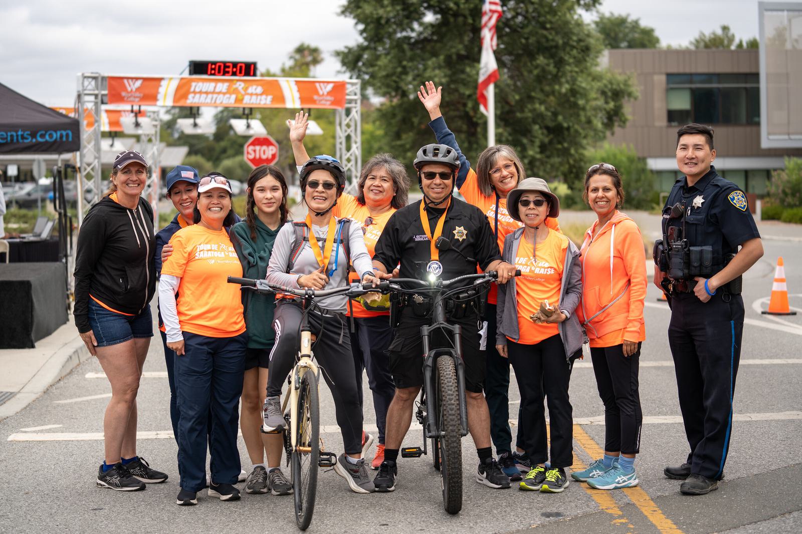 A smiling group of people in bright orange shirts pose at a race finish line, with one man on a bike and a police officer standing beside them. Several wear medals, and one woman waves enthusiastically.