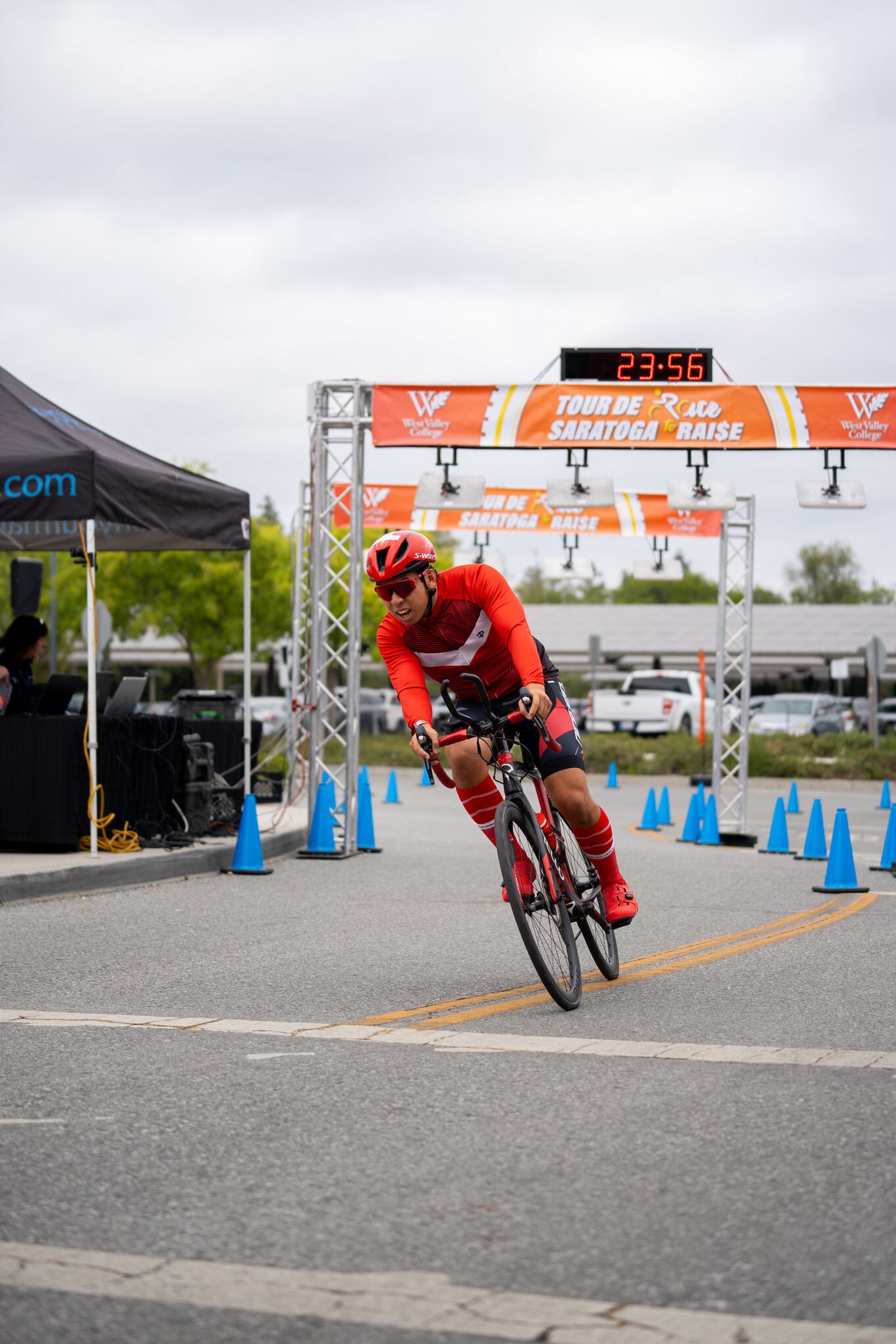 A cyclist in red gear rounds a corner during a race, with a "Tour de Saratoga Race" banner, digital timer, and blue cones visible in the background on a cloudy day.