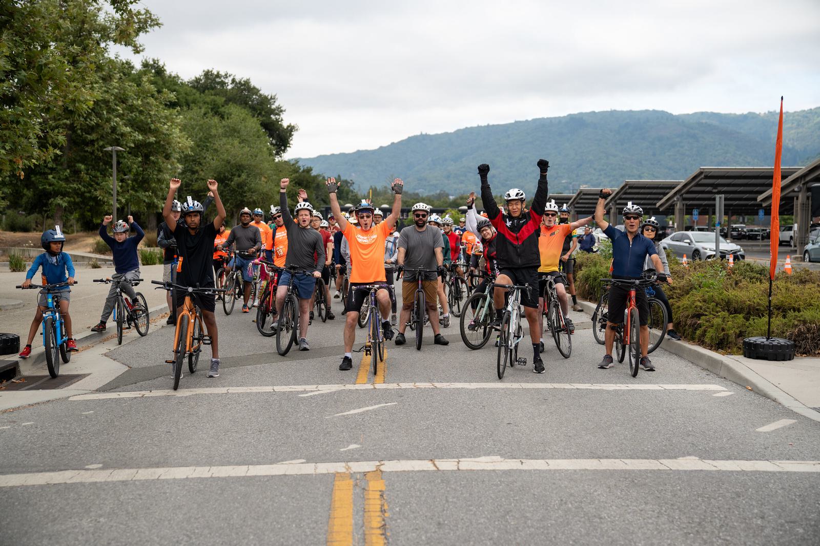 A group of cyclists, some raising their arms in celebration, pose together on a road. Trees, parked cars, and hills are visible in the background. The mood is cheerful and energetic.