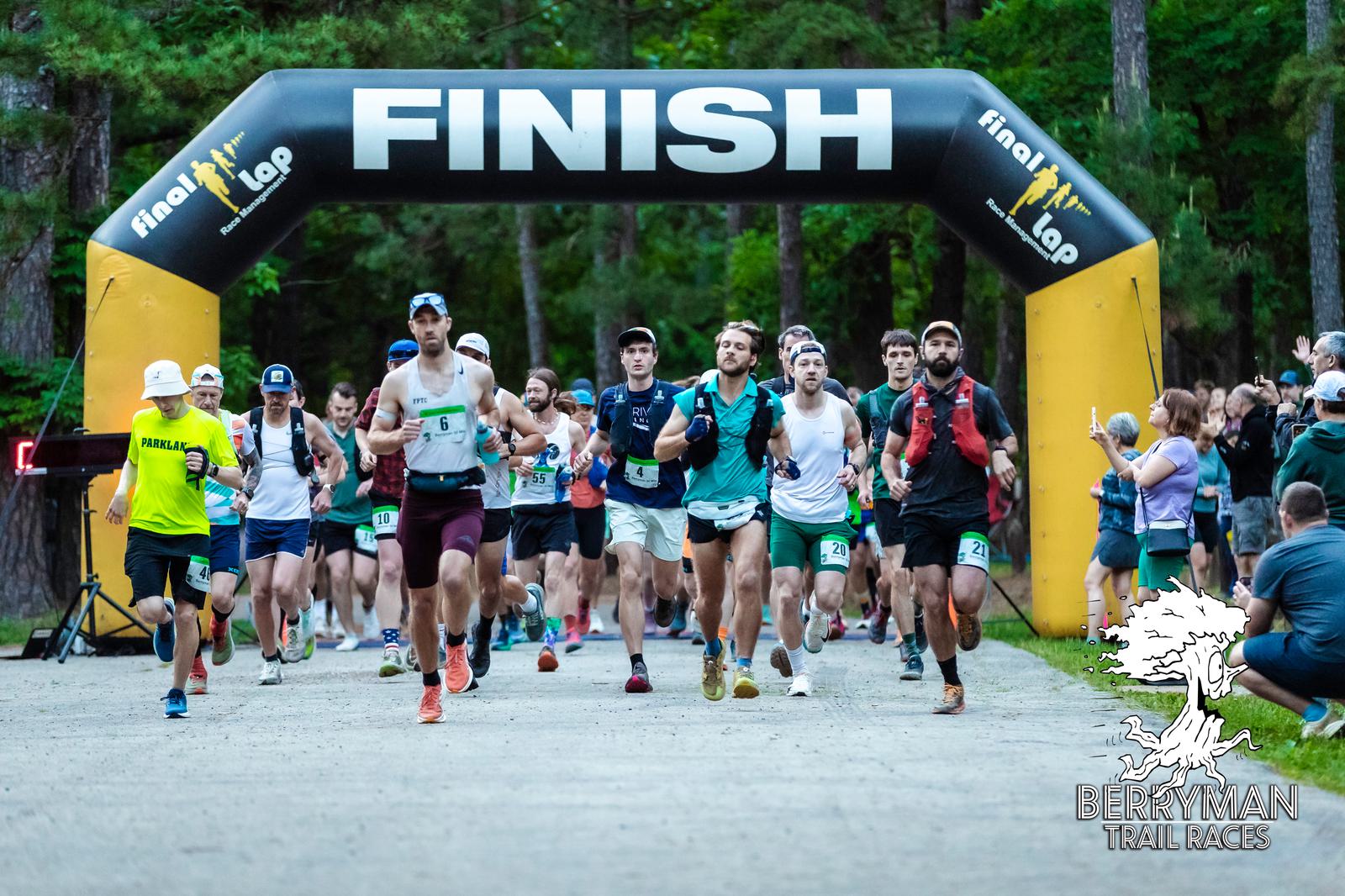 A group of runners starts a trail race under a large black and yellow "FINISH" arch in a forested area, with spectators watching. The event is labeled as "Berryman Trail Races" at the bottom right.