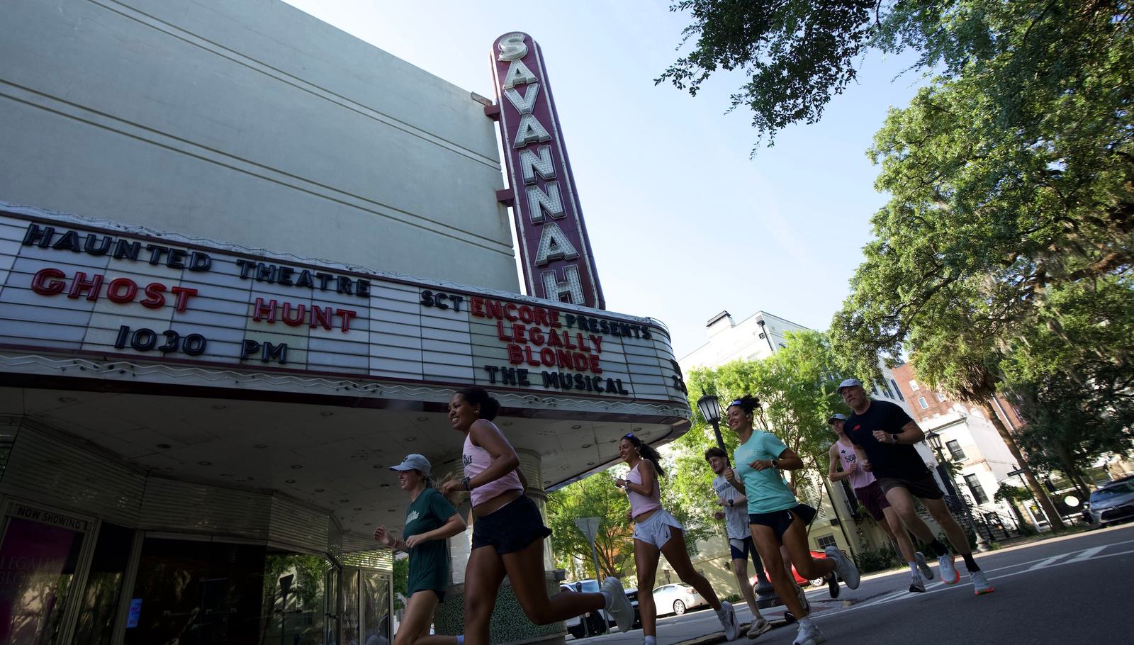 A group of people jog past the Savannah Theatre, which has a vertical neon sign and a marquee advertising a ghost hunt and "Legally Blonde The Musical." Trees and city buildings are visible in the background.