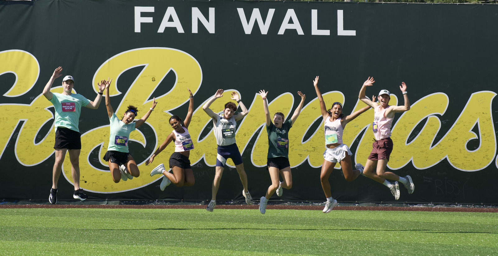 Seven people are jumping in the air with their arms raised and smiling, in front of a large wall sign that reads “Fan Wall” and “Go Bananas” on a sports field.