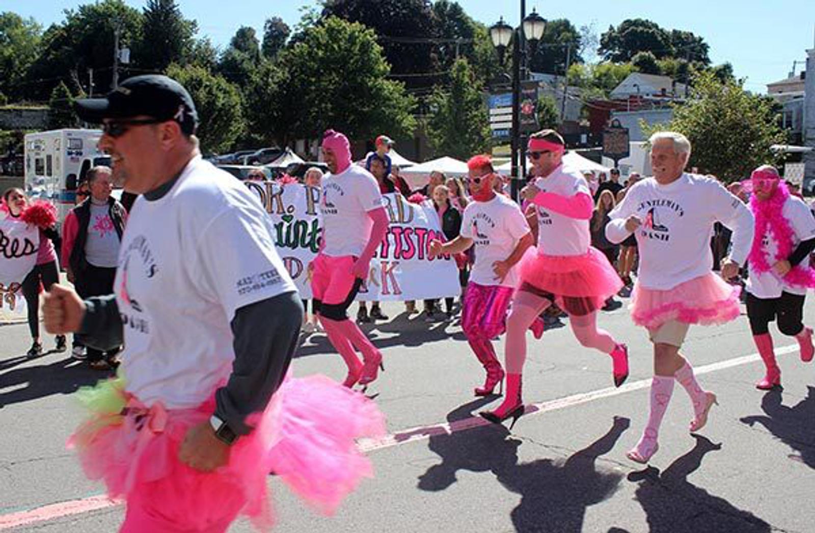 A group of people wearing pink tutus and white shirts run down a street during a parade or charity event, with others cheering and holding supportive banners in the background.