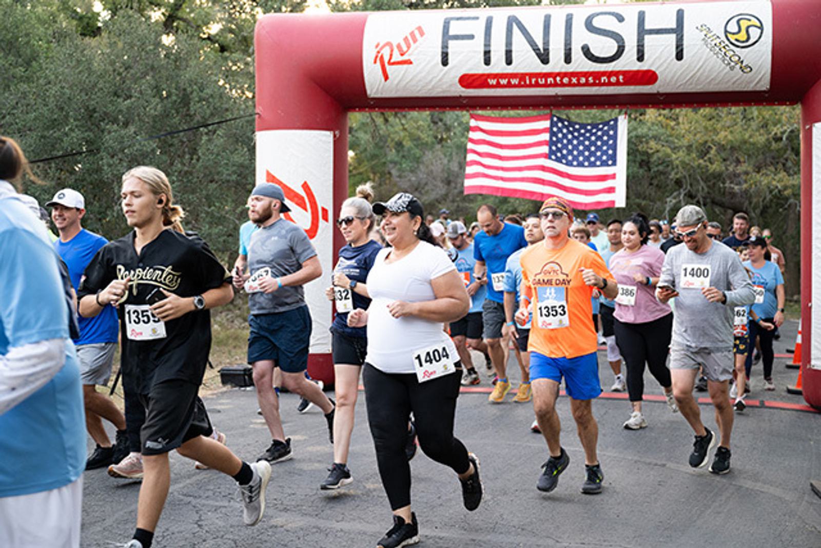 A group of runners, each wearing numbered bibs, cross the finish line under an inflatable arch labeled "FINISH" at an outdoor race event, with a large American flag displayed in the background.