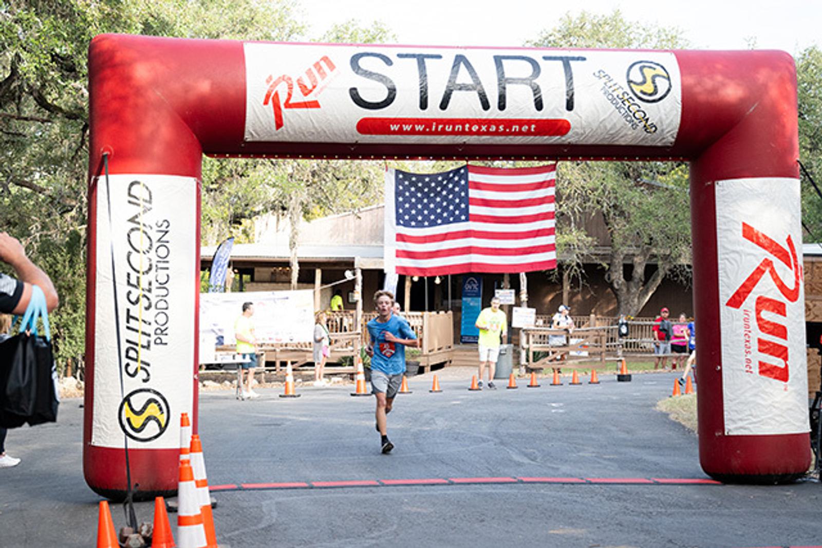 A runner crosses beneath a red and white inflatable "START" arch at a race, with an American flag and several people in the background. Orange cones line the road under trees and clear skies.