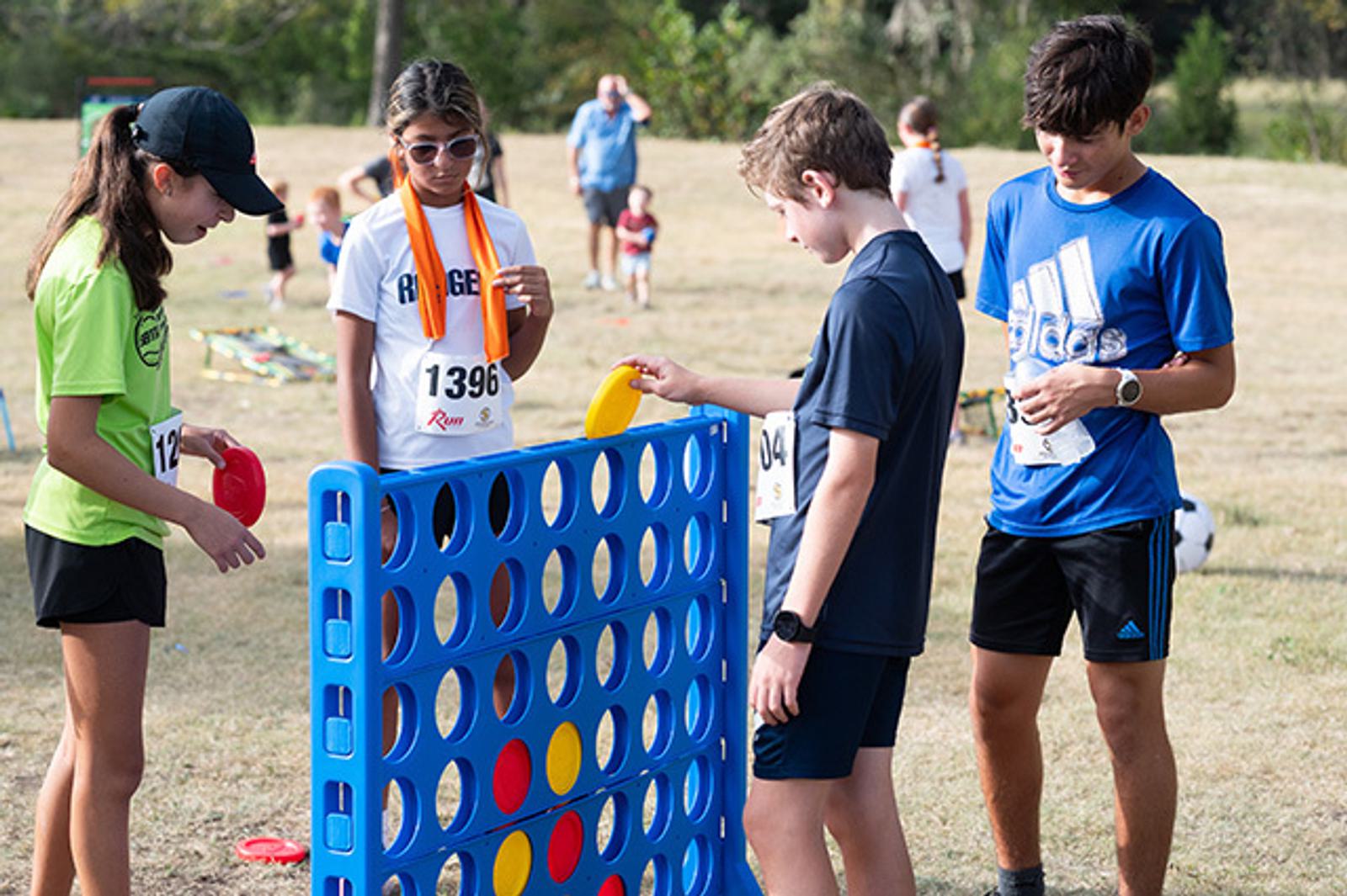 Four kids stand outdoors playing a large Connect Four game on a grassy field. They wear casual clothes and race bibs, suggesting they're at a community event or race. Other people and children are visible in the background.