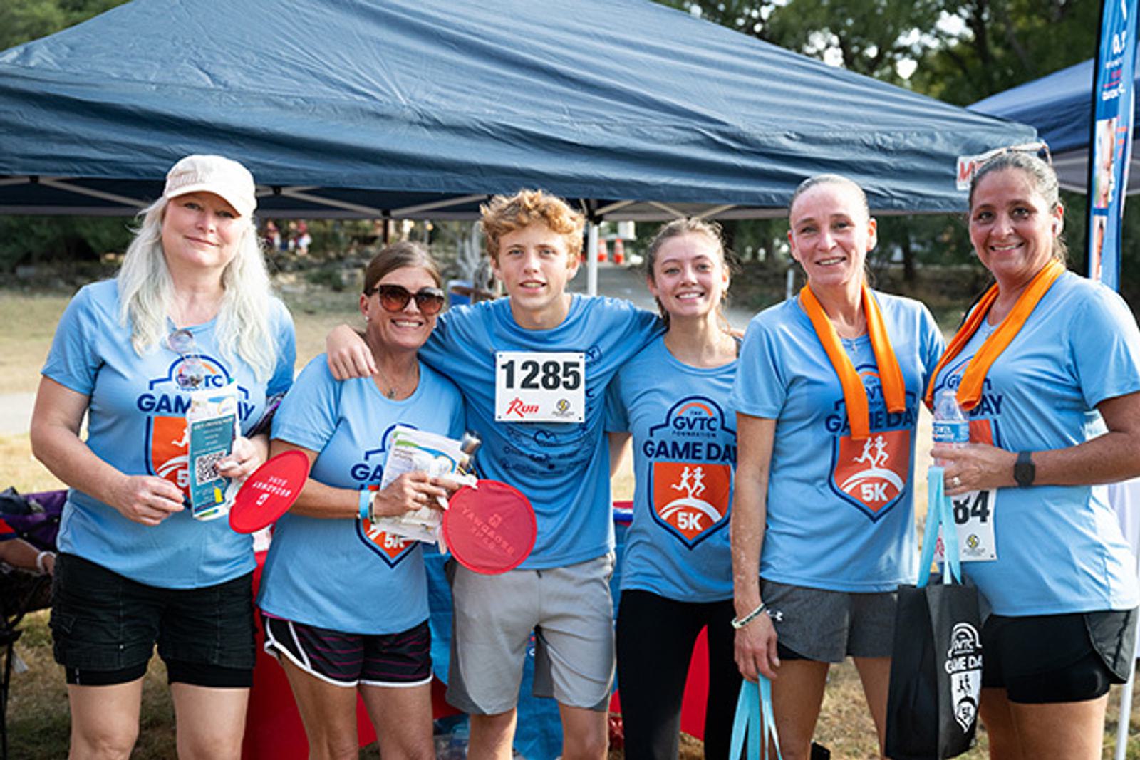 Six people in blue "Game Day 5K" shirts stand smiling under a canopy. Three women wear medals, one person holds frisbees, and a runner in the center wears bib number 1285. It's a sunny outdoor event.