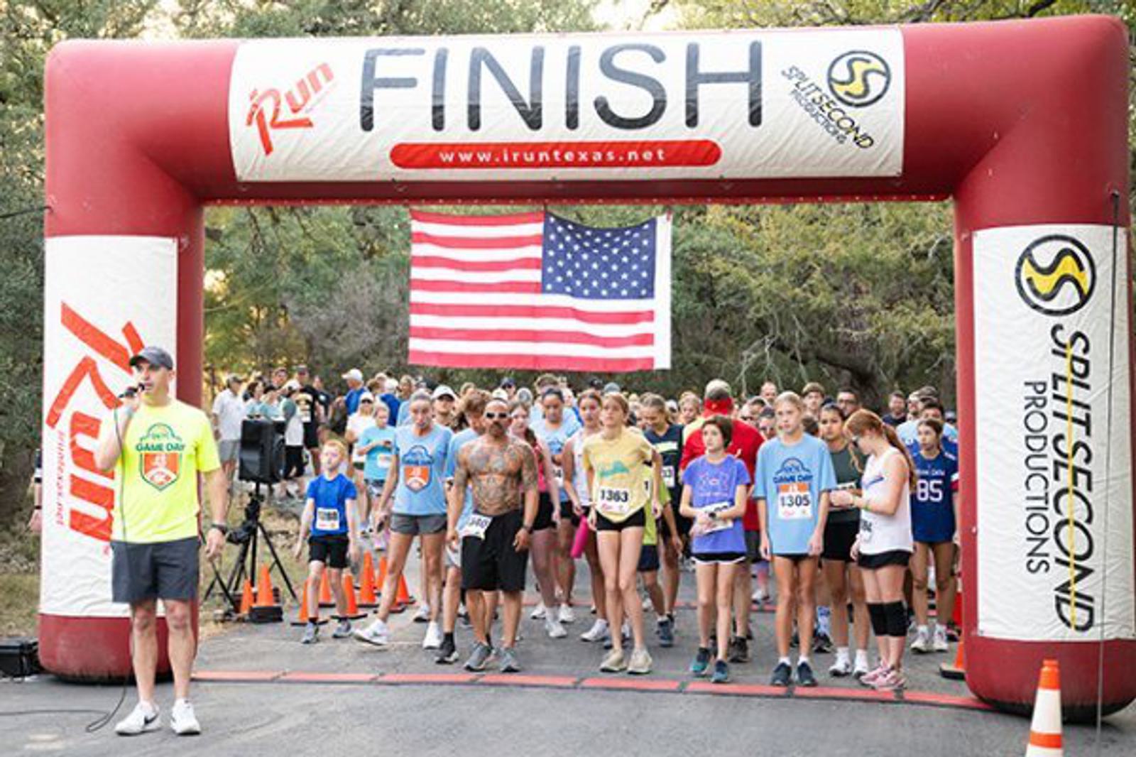 A group of runners gathers at the starting line under a large inflatable arch labeled "FINISH" with an American flag hanging behind them. A man with a microphone stands to the side, addressing the participants.