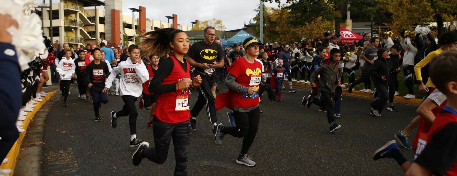 A group of people, including children and adults, run in a road race. Some runners wear costumes, such as Batman and Wonder Woman. Spectators and cheerleaders line the street, cheering them on. Trees and buildings are in the background.