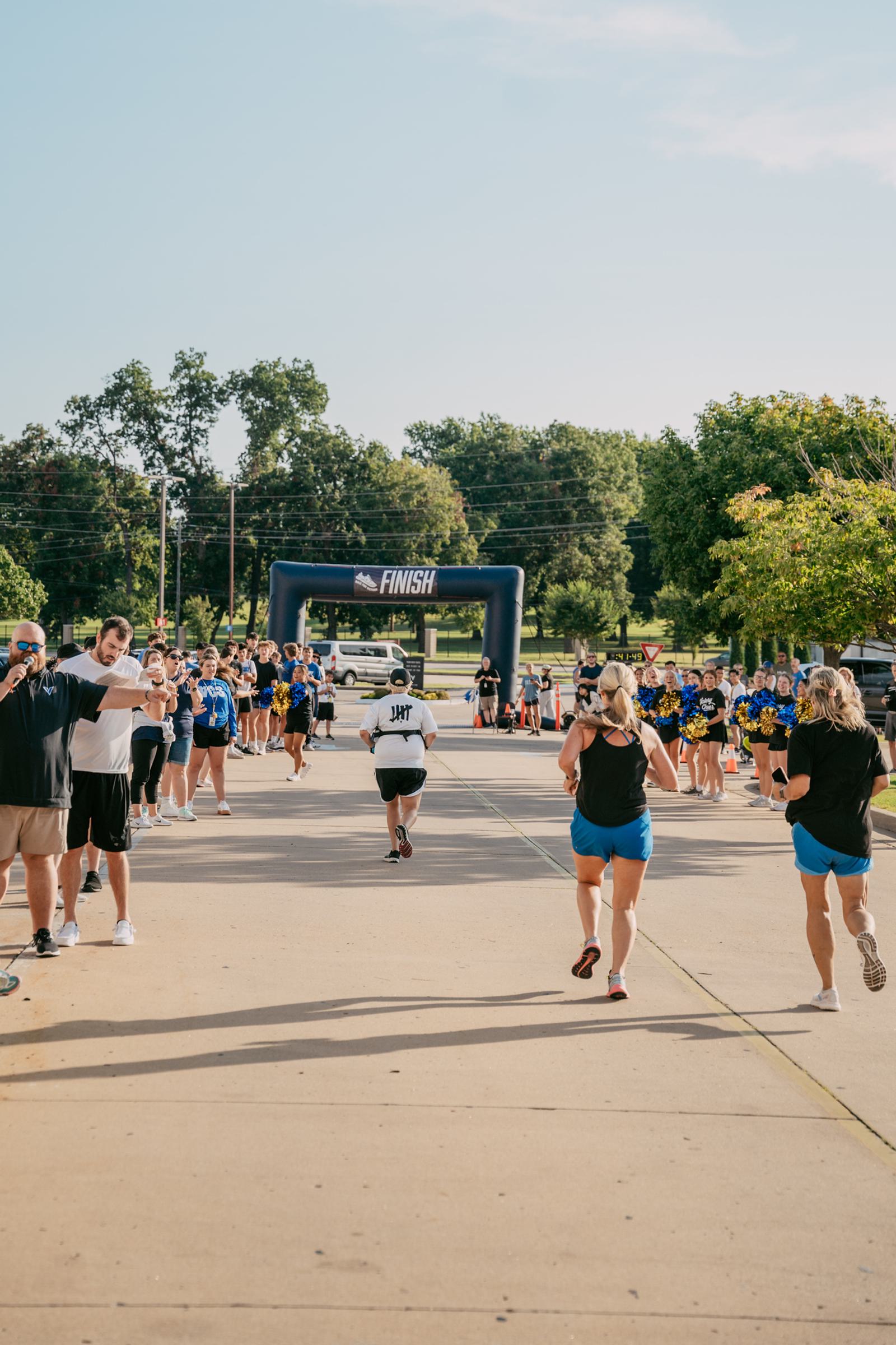 Runners head toward a blue finish line arch, cheered on by spectators and supporters lining both sides of a wide, sunlit path surrounded by green trees.