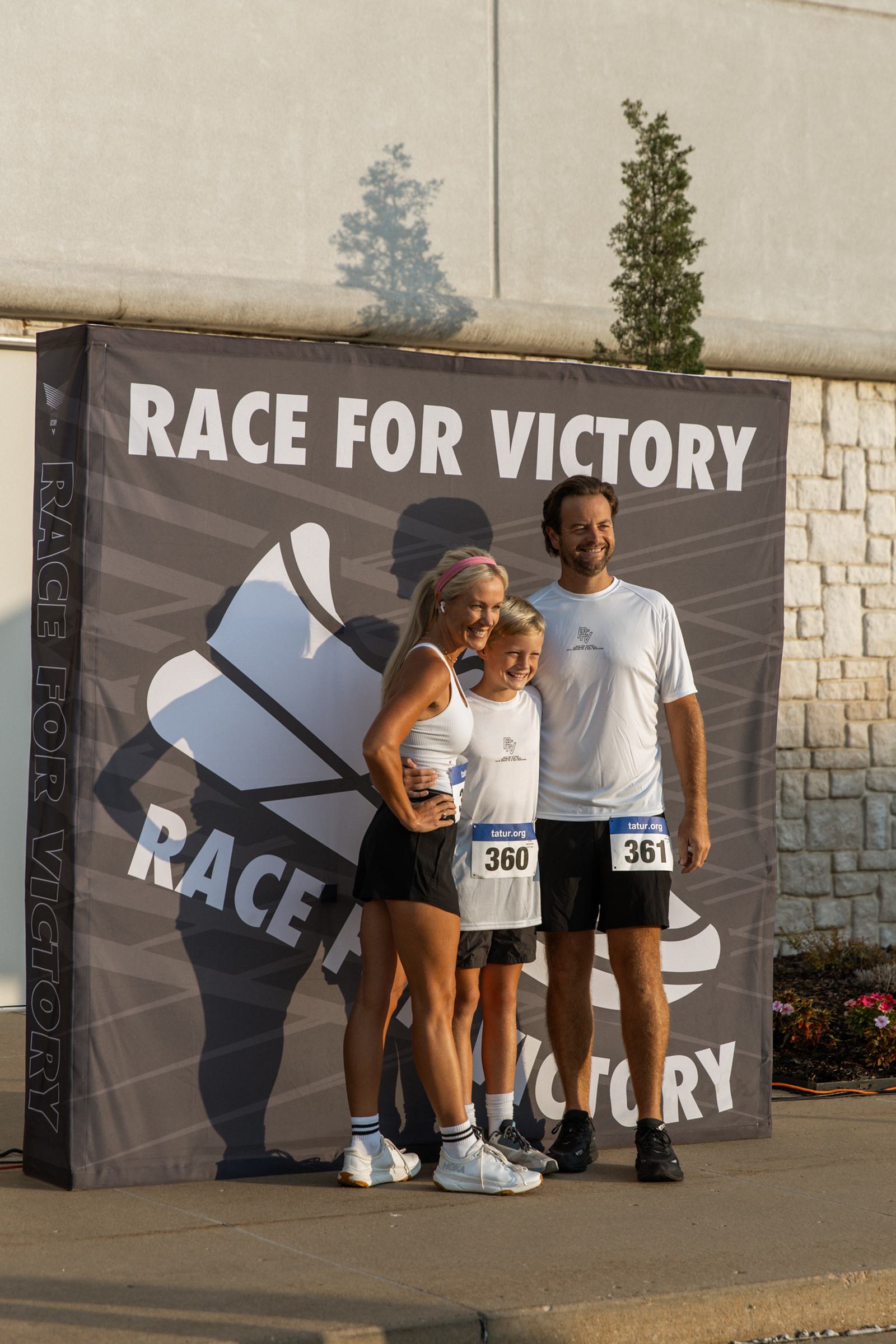 A smiling family of three in athletic gear and race bibs poses in front of a "Race for Victory" banner outside on a sunny day.