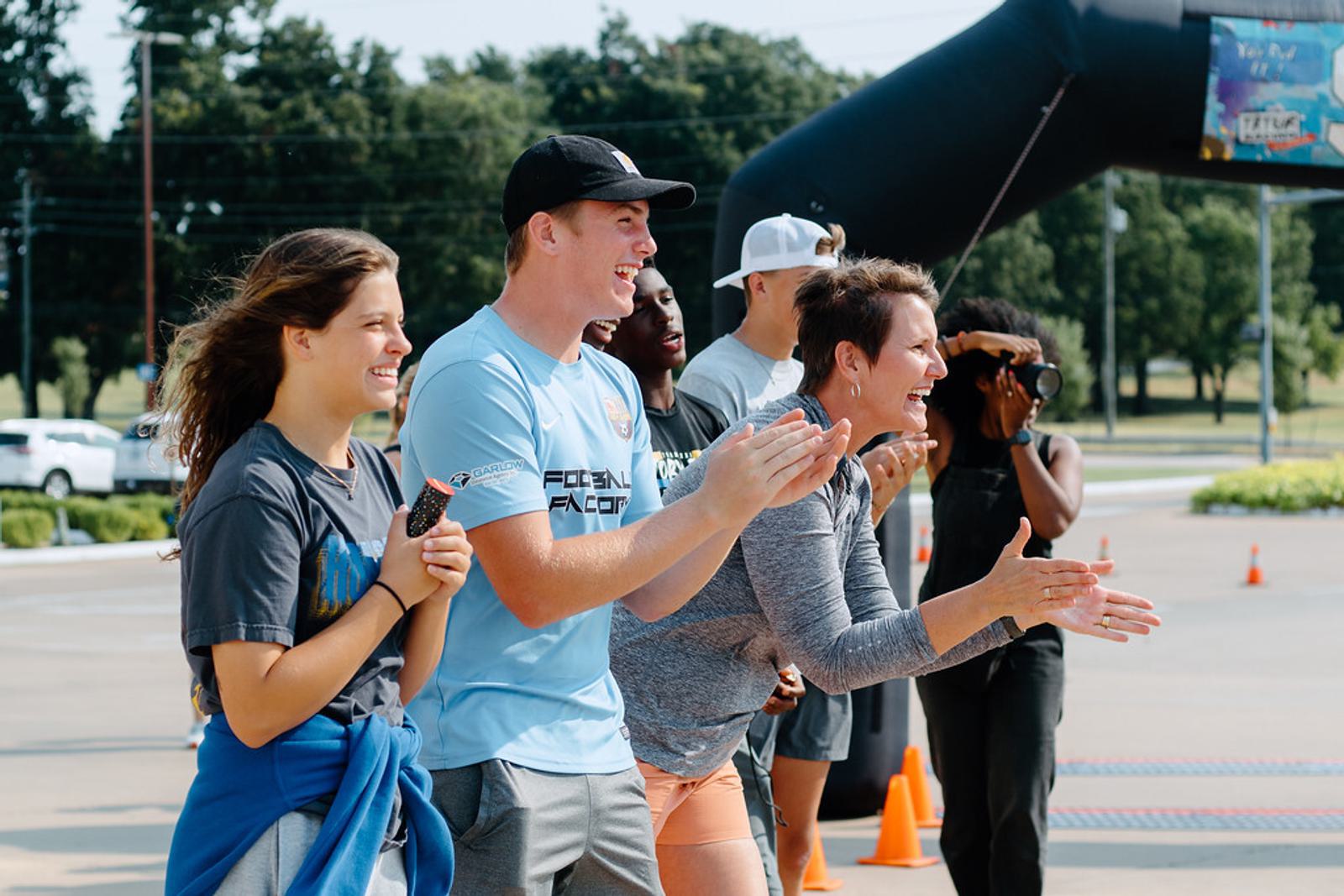 A group of people stand outdoors, smiling and clapping enthusiastically. They appear to be cheering for an event. Orange cones and an inflatable arch are visible in the background.