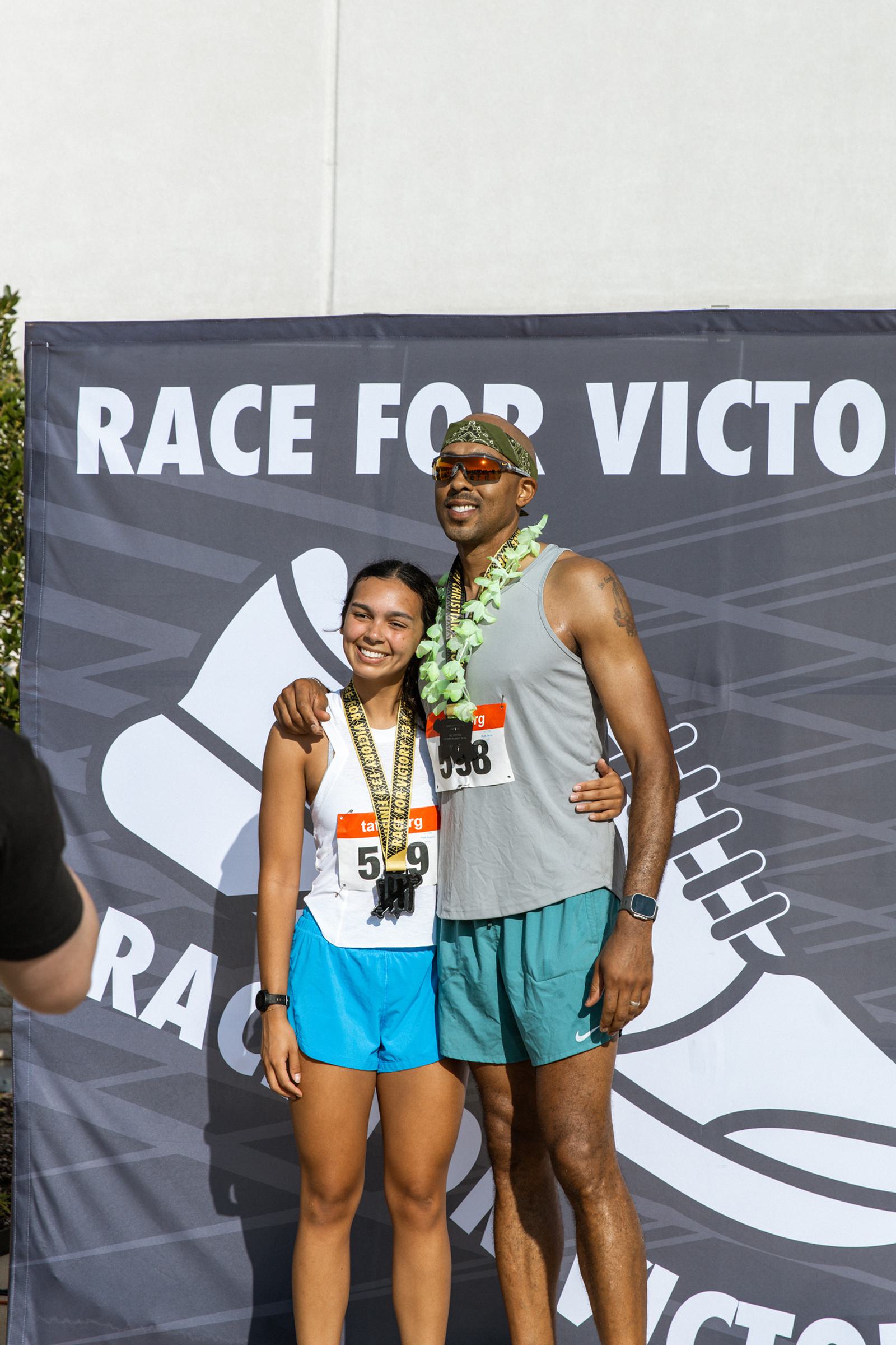 Two race participants in athletic gear stand smiling with medals around their necks in front of a "Race for Victory" banner. They appear happy and proud after completing the event.