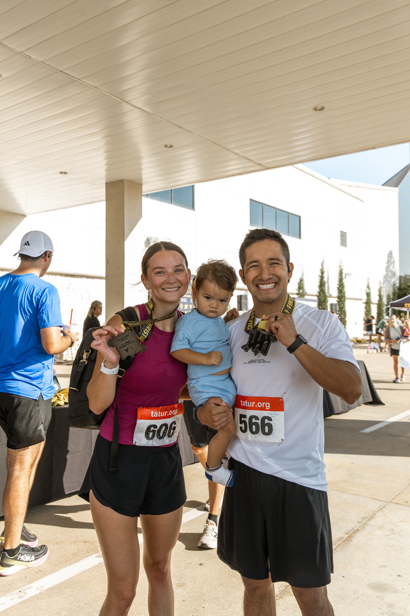 A smiling woman and man wearing race medals and bibs pose with a young child outside a building after a race event. The woman and man hold up their medals, and other participants are visible in the background.