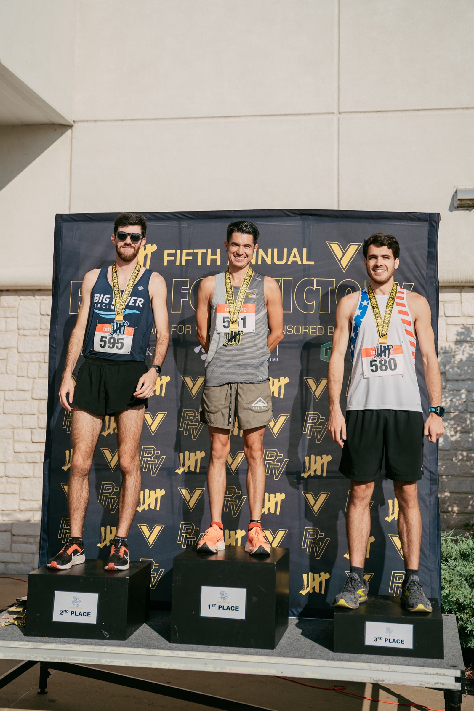 Three male runners stand on a podium with medals around their necks, celebrating their 1st, 2nd, and 3rd place wins at the Fifth Annual Big Victory event, with a branded backdrop behind them.