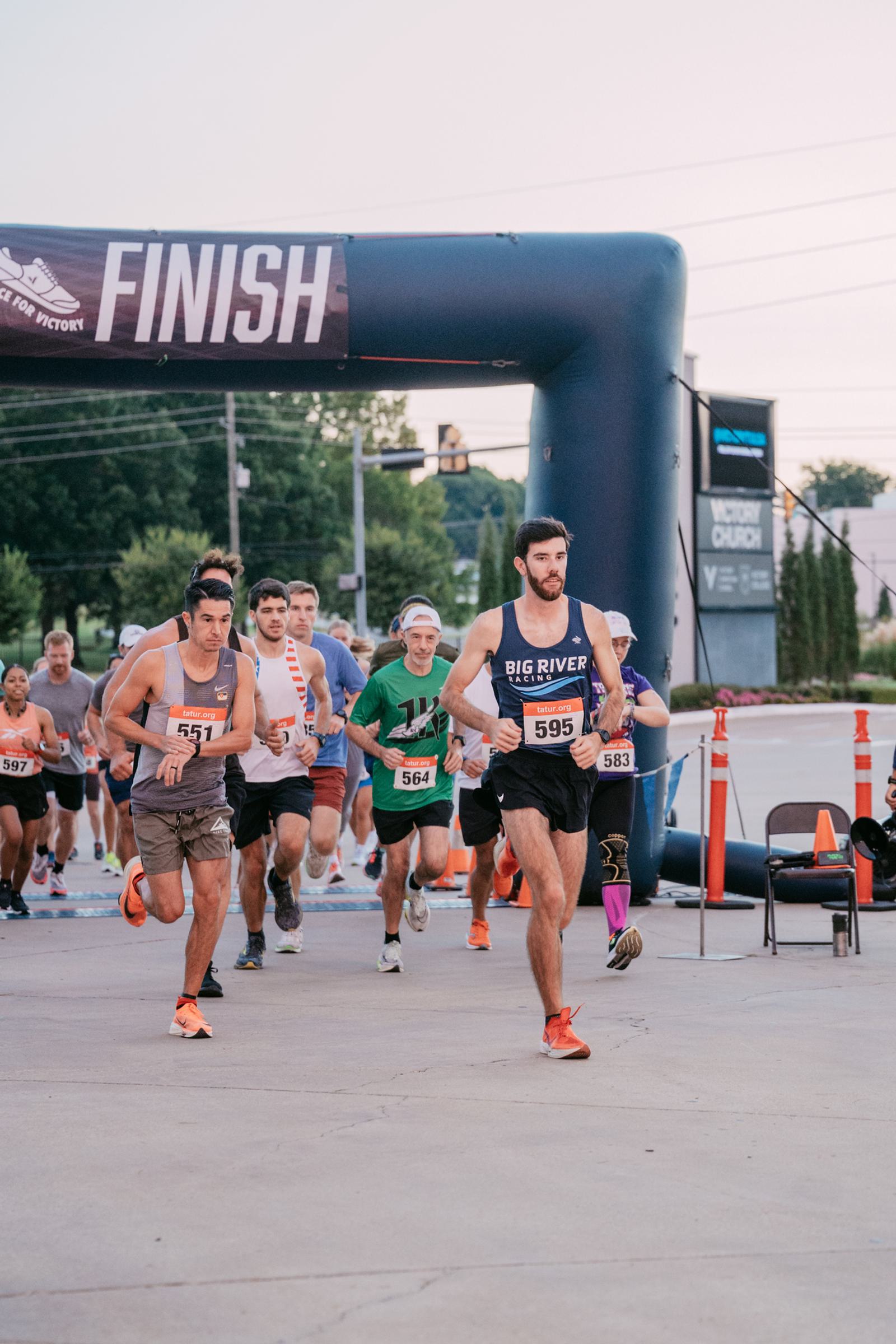 Runners in athletic gear approach the finish line during a road race, with a large inflatable "FINISH" arch overhead and race numbers pinned to their shirts. Spectators and traffic cones are visible in the background.