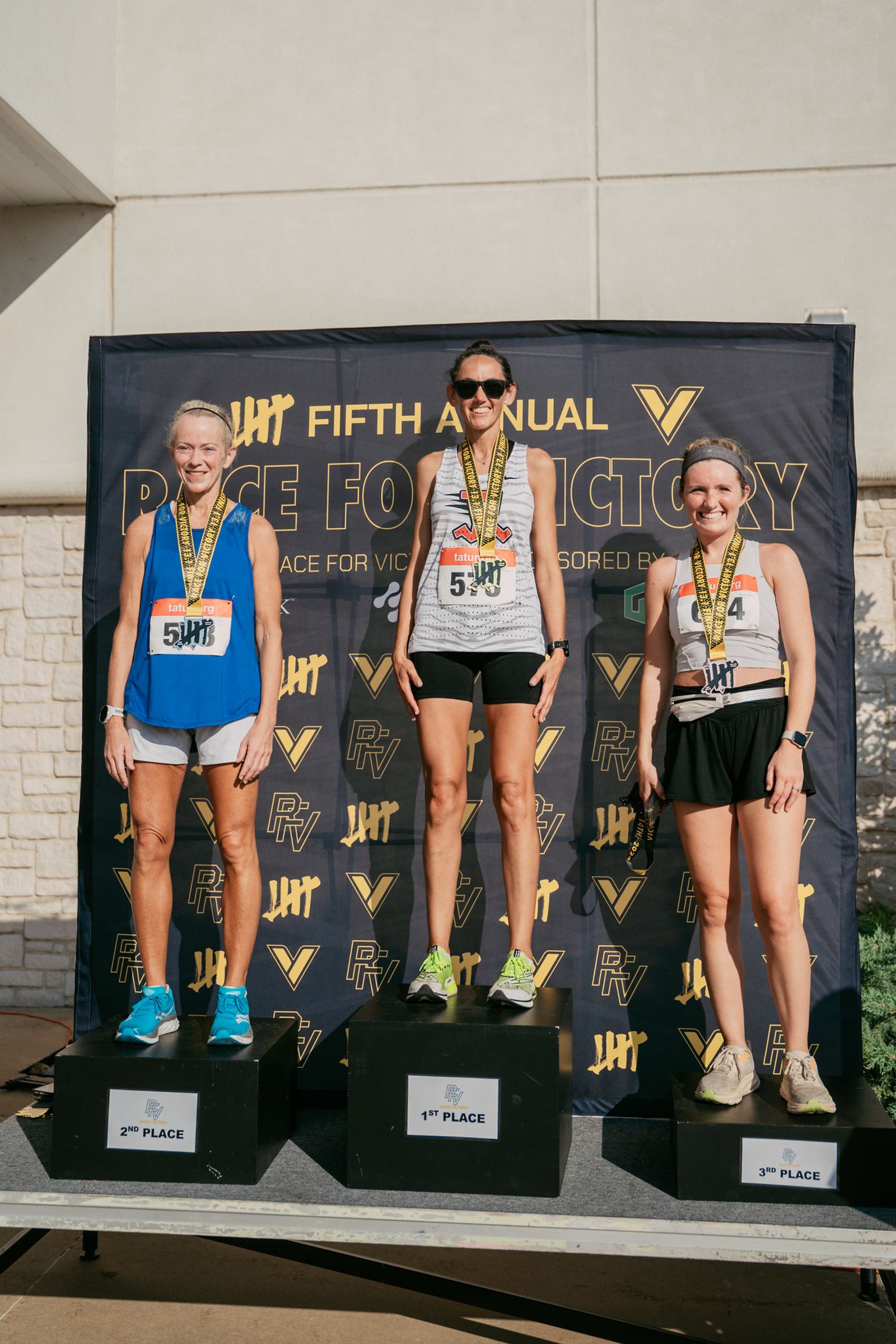 Three female runners stand on podiums for 1st, 2nd, and 3rd place at a race event. They smile and wear race bibs and medals, with a "Race for Victory" banner in the background.