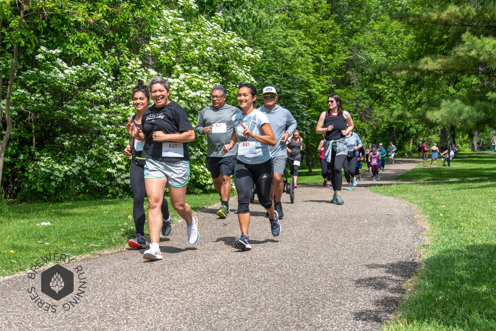 A group of people jogging and smiling on a paved path through a green park, with trees and shrubs in the background. More runners are visible further along the path. A logo for the Brewery Running Series is in the corner.