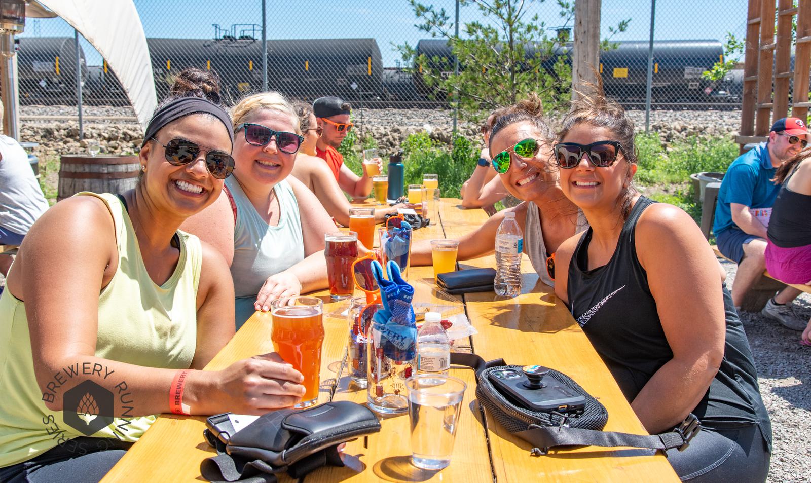 Four women sit at a sunny outdoor table, smiling and holding drinks. Other people socialize in the background beside a train track and a chain-link fence. A logo says “Brewery Run.” Drinks and sunglasses are on the table.
