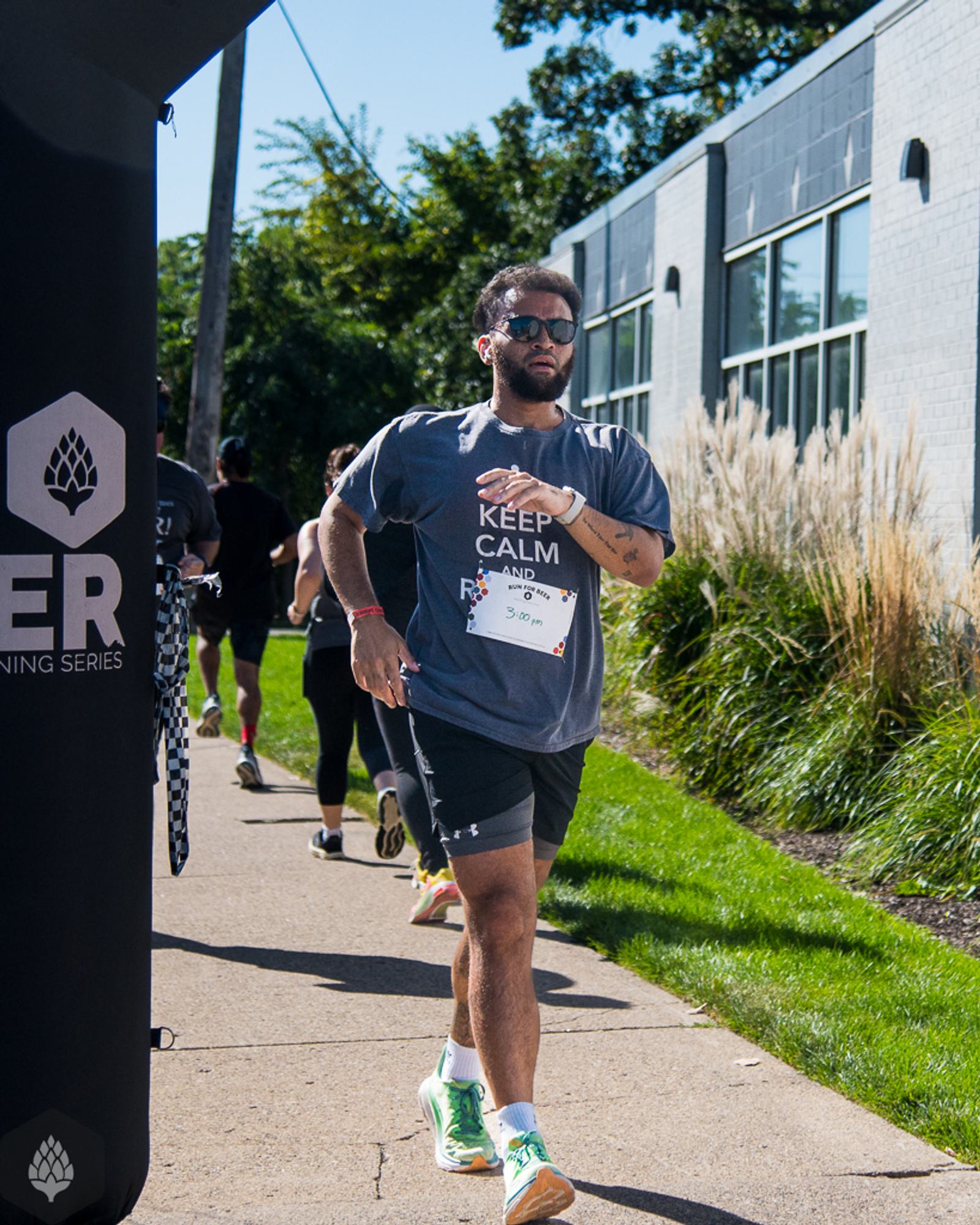 A man wearing sunglasses and a "Keep Calm" T-shirt runs in a race on a sunny day, passing an event banner near a modern building and tall grass, with other runners visible in the background.