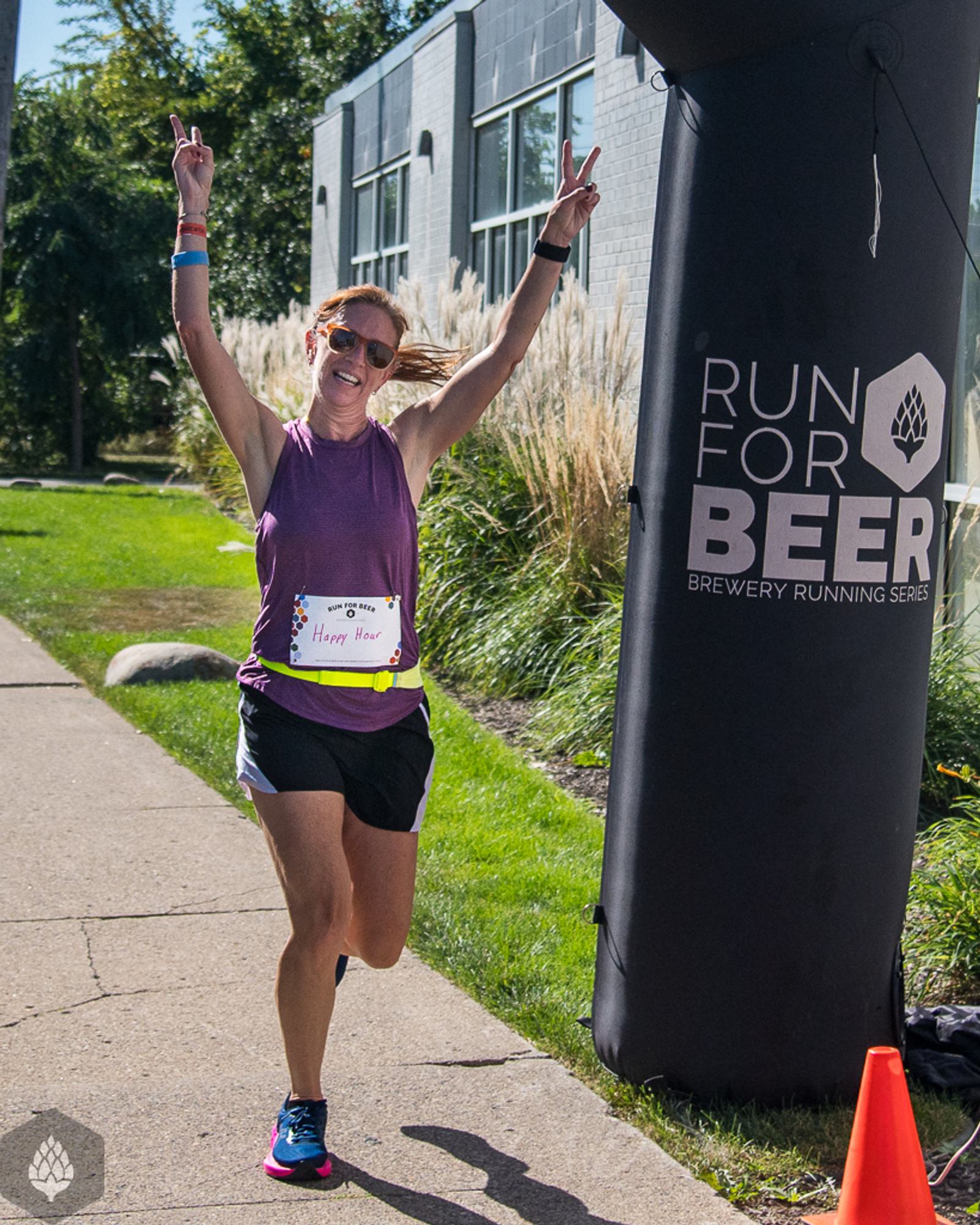 A woman in running gear smiles and raises both arms in celebration as she crosses the finish line at a "Run For Beer" event on a sunny day.