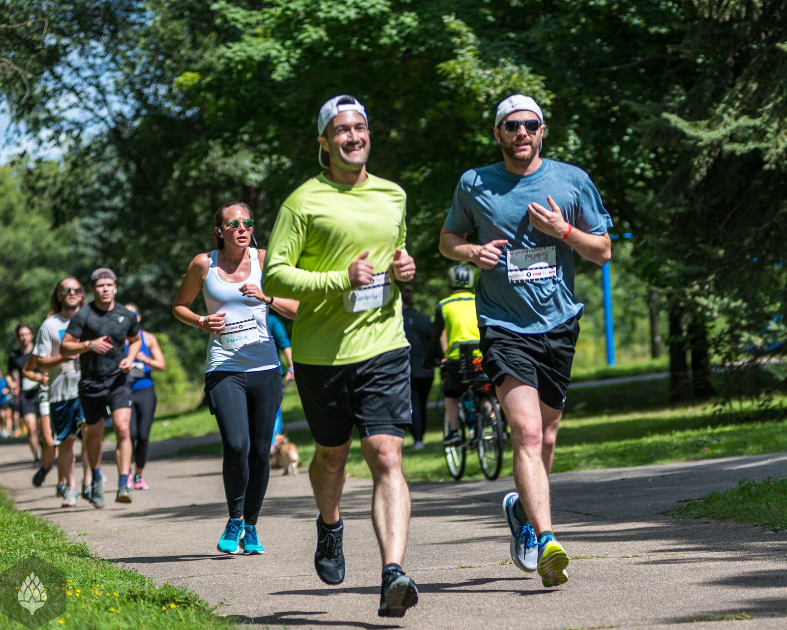 A group of people jogging on a sunny day in a park, surrounded by green trees. The runners are smiling and appear to be participating in a race or fitness event.