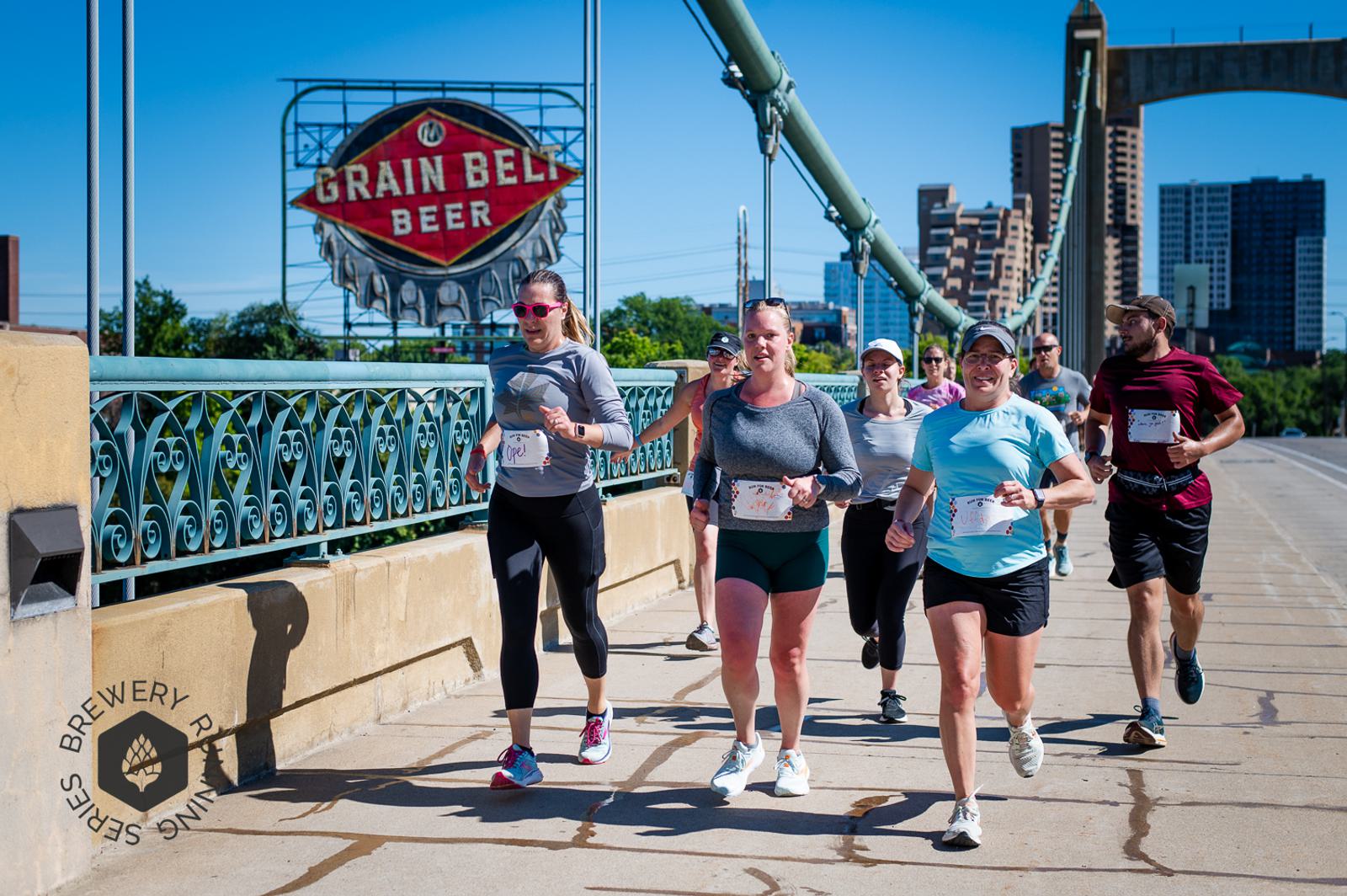 A group of runners participate in a race on a sunny day, crossing a bridge with a large Grain Belt Beer sign and city buildings visible in the background. The runners are smiling and wearing race bibs.